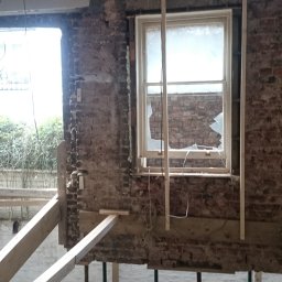 BEBEK, BEBEK MAN BUŁKA I SPÓŁKA DARIUSZ LENART - Wnętrze w trakcie remontu: odsłonięta cegła, uszkodzone okno z rozbitym szkłem, drewniane podpory i fragment sufitu z płyt gipsowych. Widoczne ślady prac budowlanych.