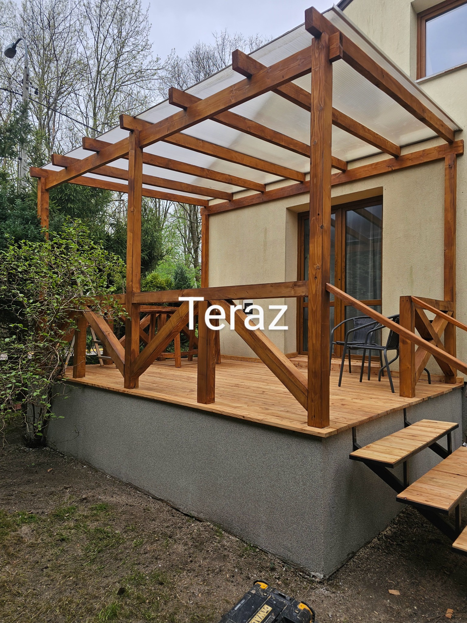 Zadaszony, drewniany taras z balustradą i schodami, przylegający do budynku. Widoczne krzesła i stół. Na ziemi leży skrzynka narzędziowa.