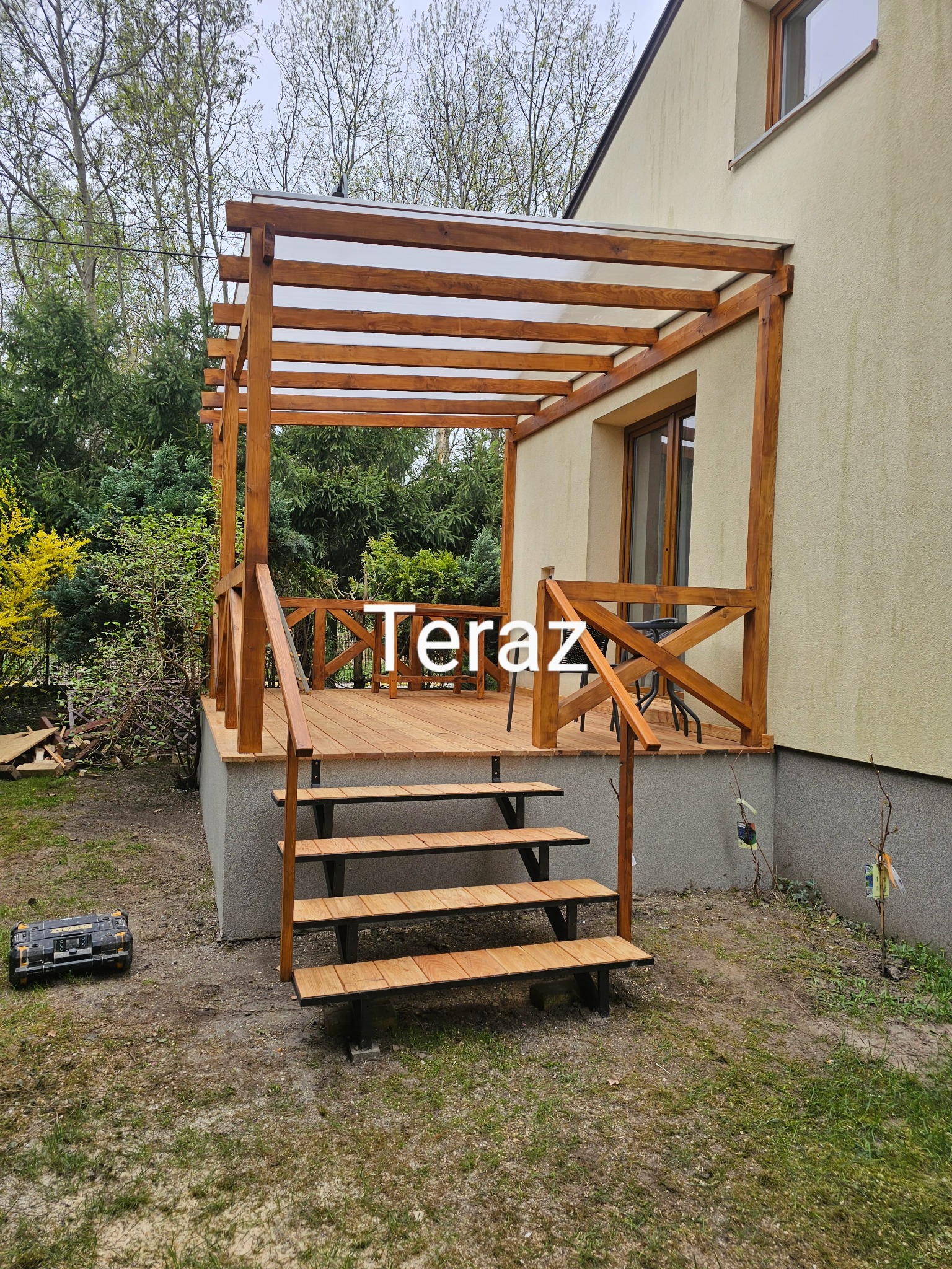 Drewniana, zadaszona weranda z balustradą i schodami, przylegająca do domu. Widoczna skrzynka narzędziowa obok schodów. Całość w ciepłych odcieniach brązu i beżu.