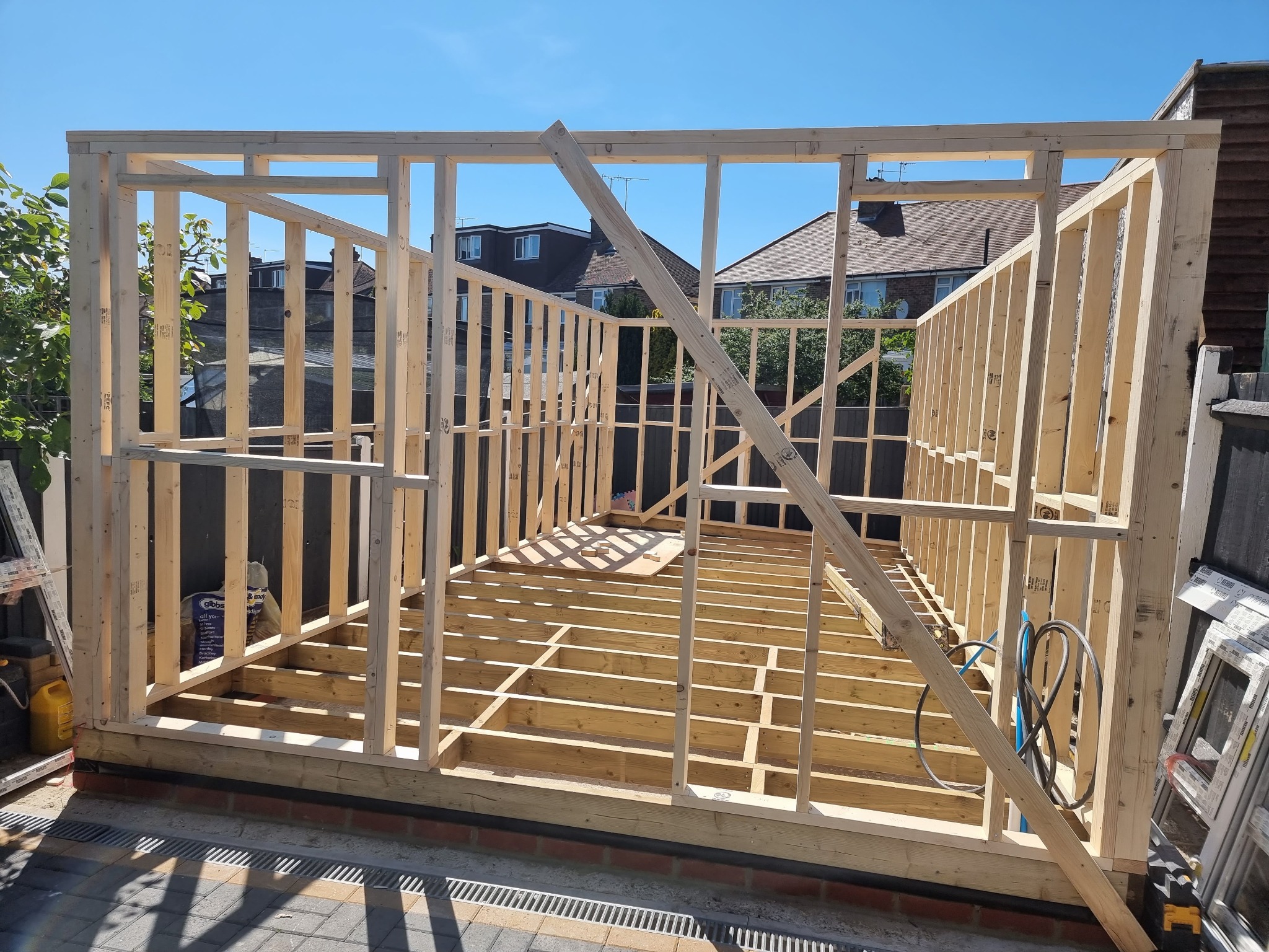 Konstrukcja szkieletowa budynku w trakcie budowy, widoczne drewniane belki i słupy, fundament z cegieł, otoczenie z zielenią i domami w tle, słoneczny dzień.