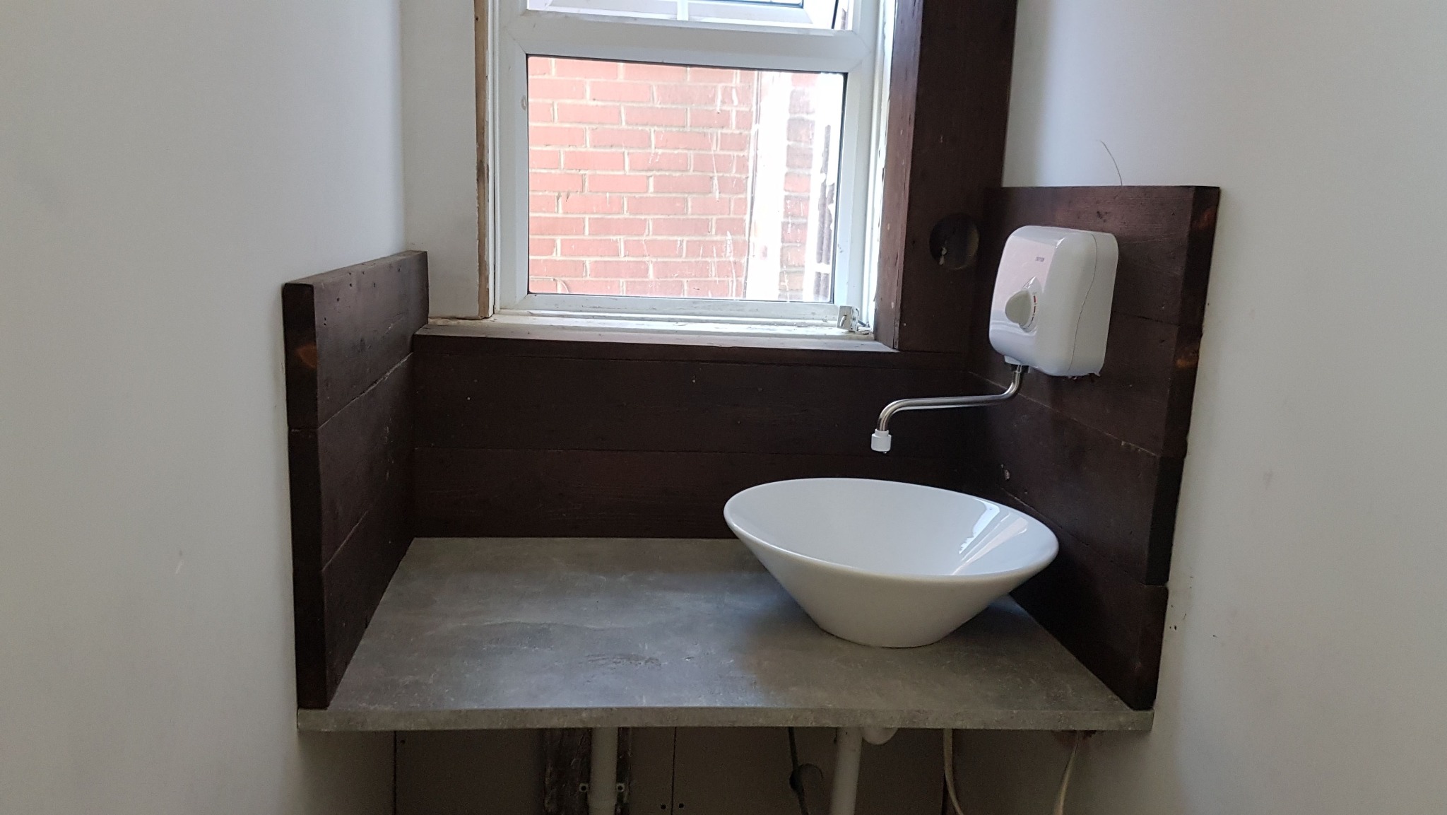 Narożna umywalka w łazience z drewnianą boazerią i blatem z szarego betonu, widok na okno z ceglanym budynkiem w tle. Minimalistyczny design.