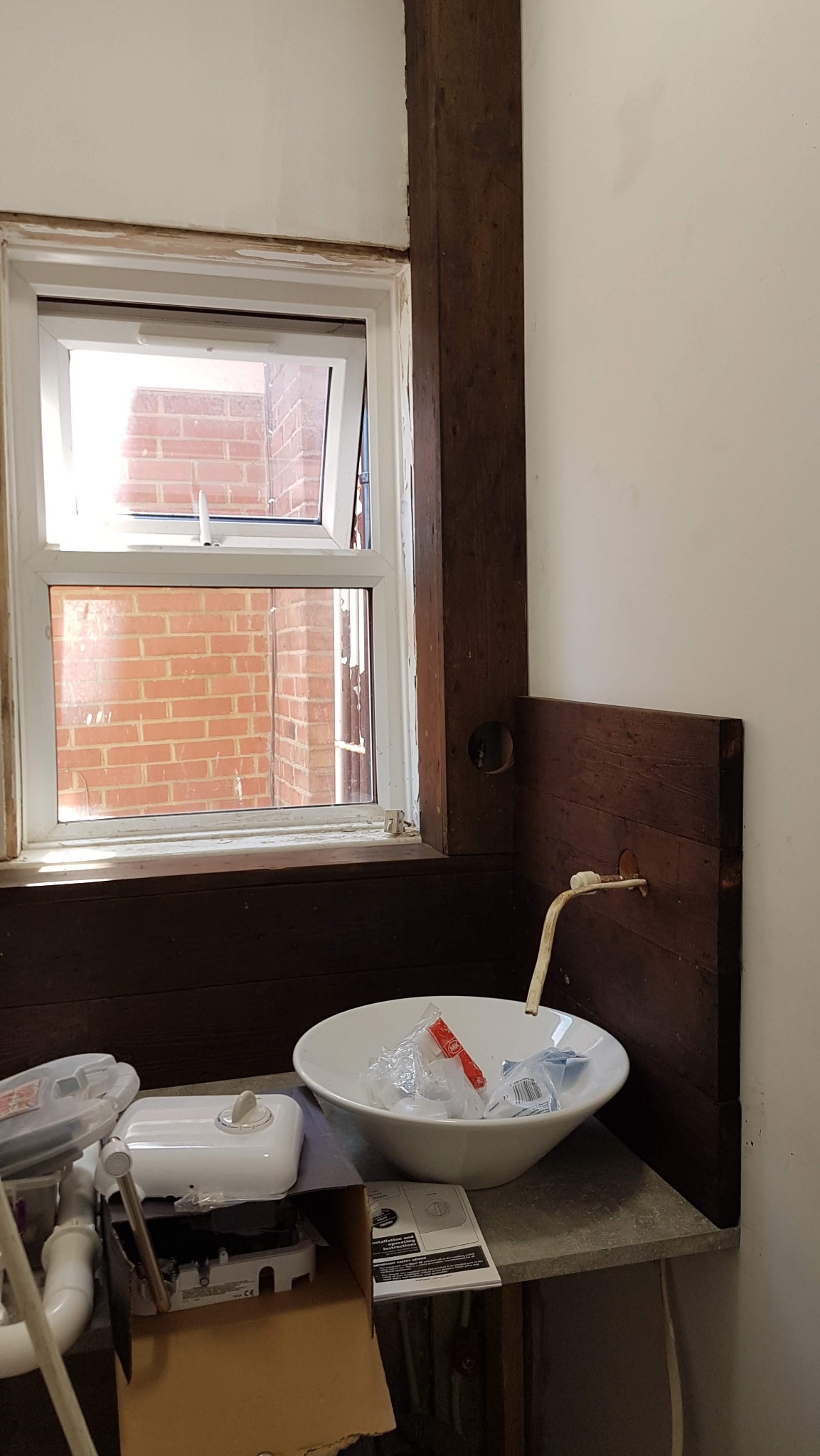 Niedokończona łazienka z umywalką w misie, drewnianą boazerią i oknem. Widoczne elementy instalacji i akcesoria montażowe. Etap prac wykończeniowych.