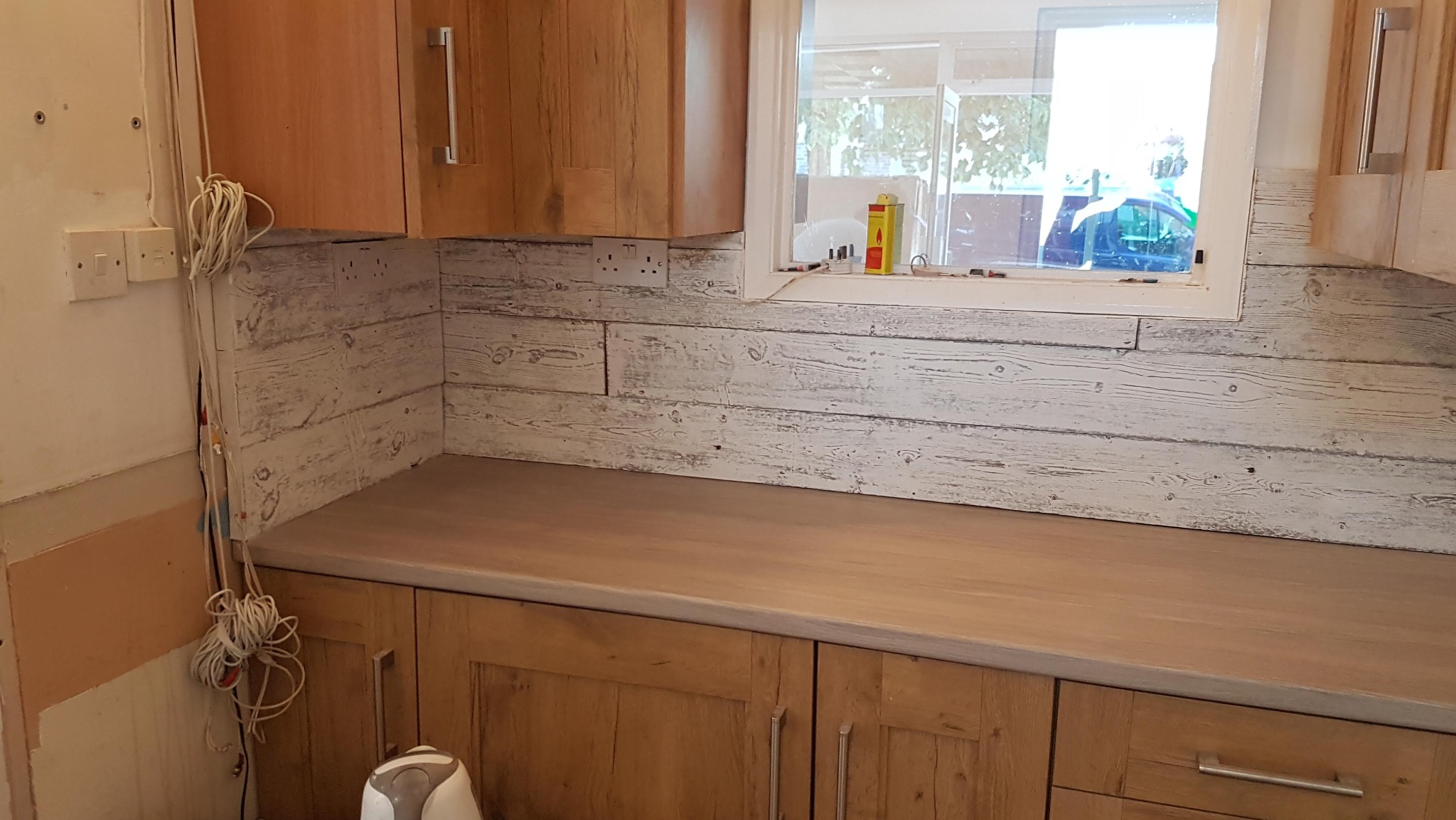 Jasna kuchnia z drewnianymi szafkami i blatem. Ściana nad blatem wyłożona panelami imitującymi bielone drewno. Widoczne gniazdka elektryczne i okno.