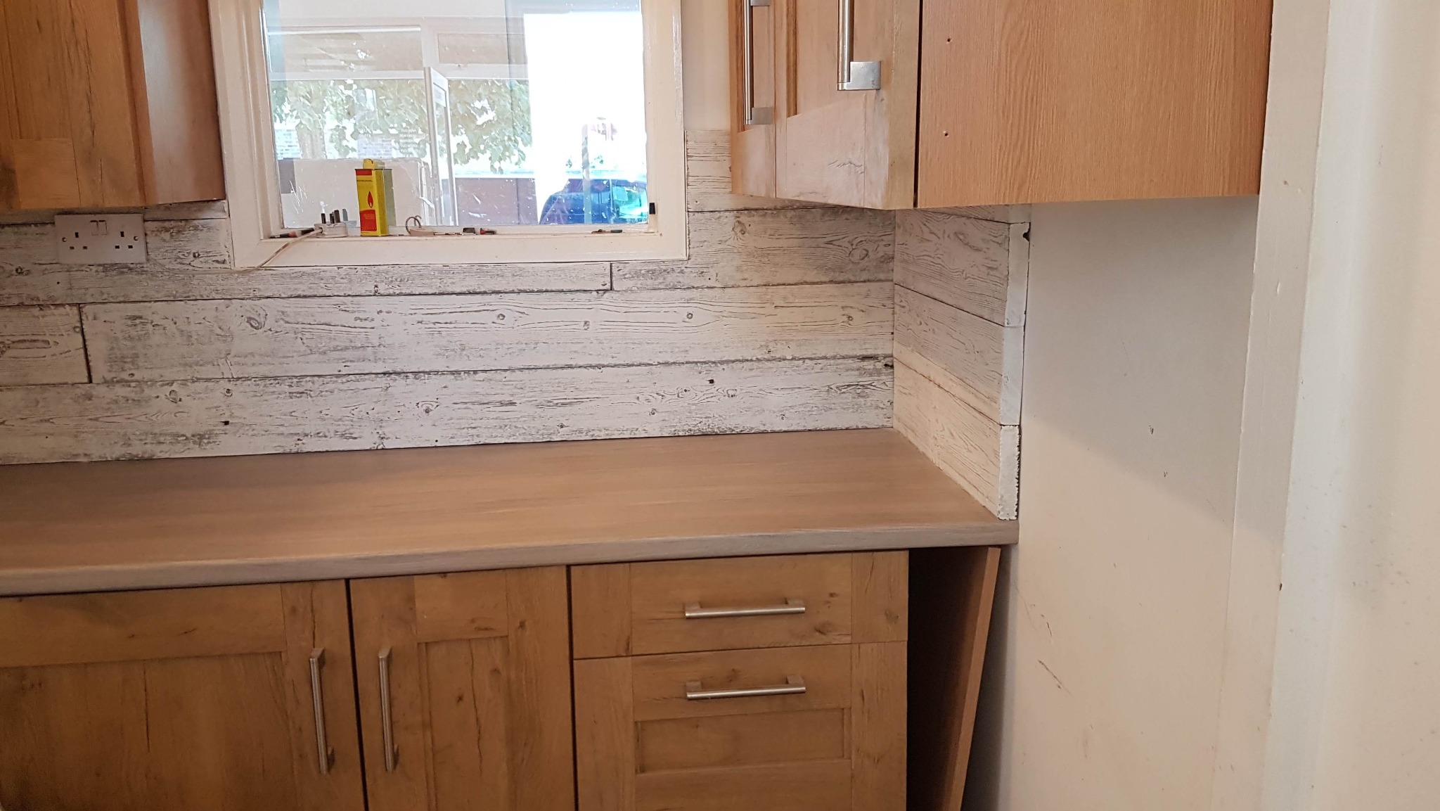 Jasna kuchnia z drewnianymi szafkami i blatem. Ściana nad blatem wykończona panelami imitującymi bielone drewno. Widoczne okno z widokiem na zieleń.