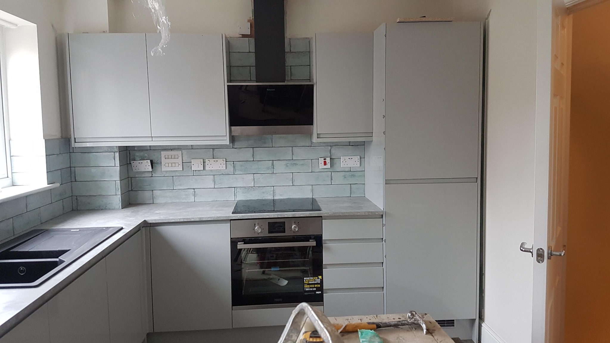 Jasna kuchnia z szarymi szafkami i turkusową mozaiką nad blatem. Widoczny piekarnik, płyta indukcyjna i okap. Na pierwszym planie narzędzia na drabinie.