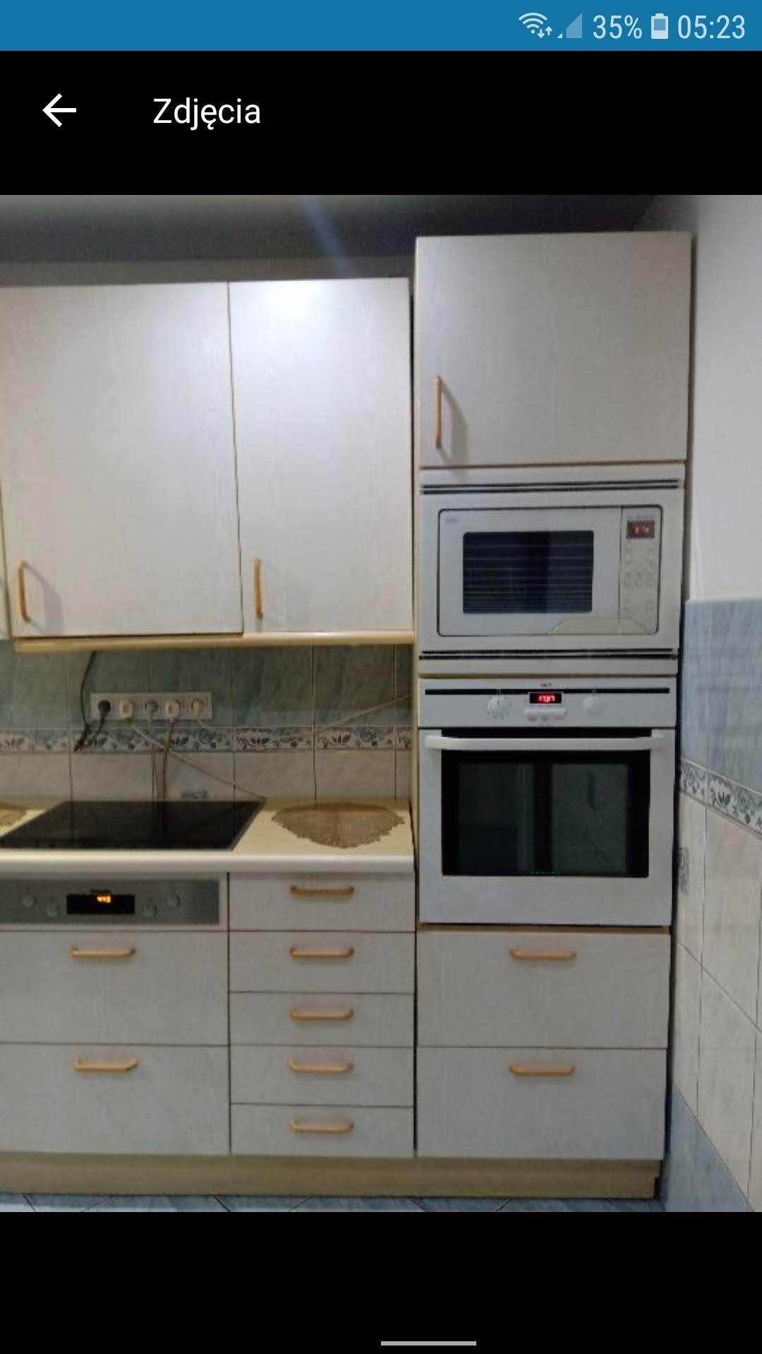 Jasna zabudowa kuchenna z drewnianymi uchwytami, piekarnikiem i mikrofalówką wkomponowanymi w szafki. Widoczna płyta indukcyjna i płytki ceramiczne na ścianie.