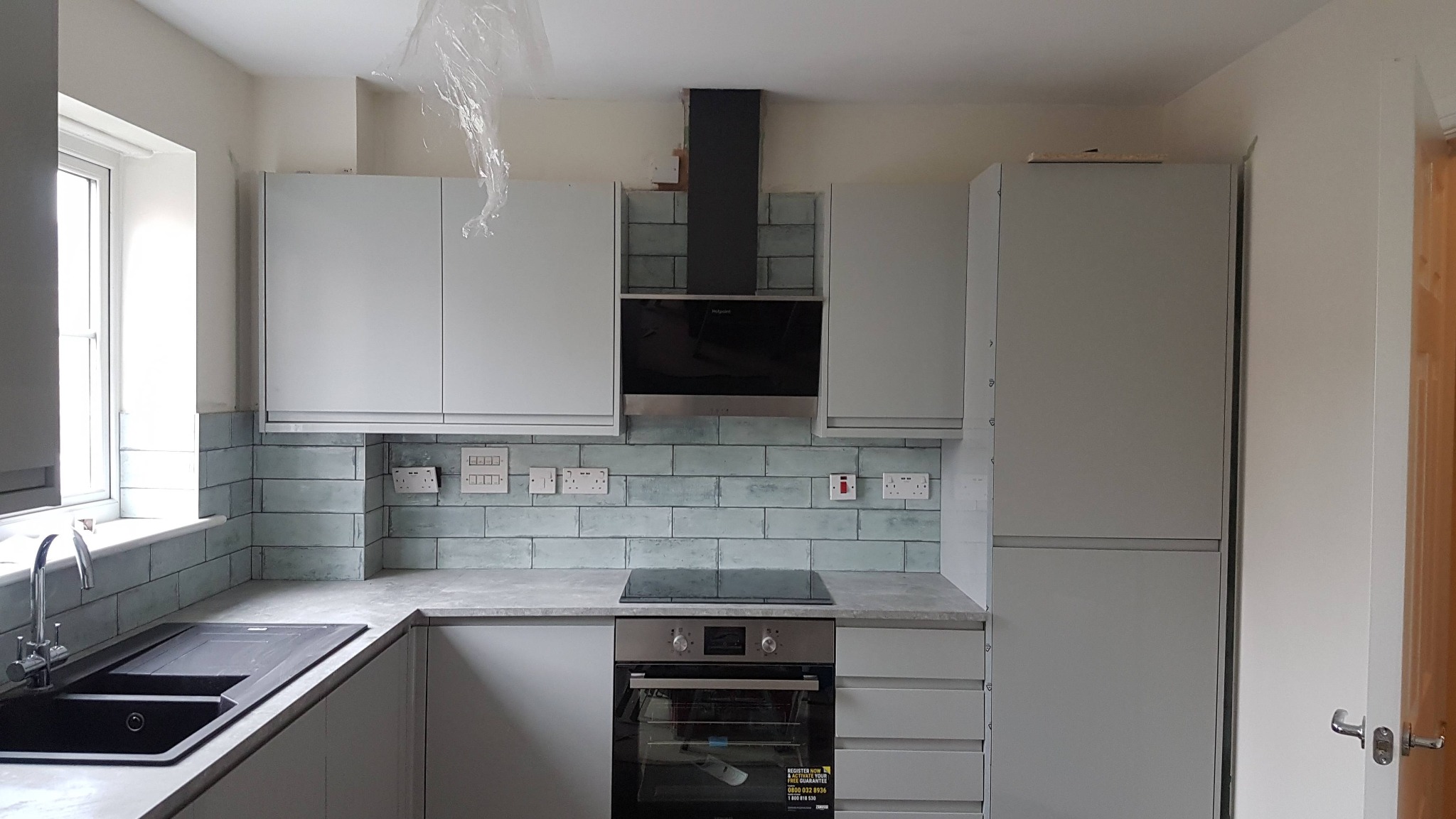 Jasna kuchnia po remoncie: szare szafki, błękitne płytki, czarny okap i zlew. Widoczne gniazdka elektryczne i płyta indukcyjna. Folia ochronna na suficie.