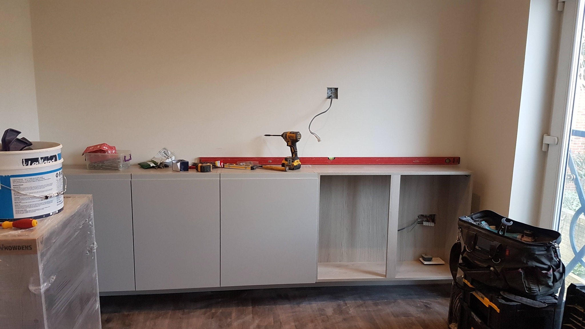Szare szafki kuchenne w trakcie montażu, poziomica, wiertarka i narzędzia na blacie, widoczne kable elektryczne w ścianie, torba narzędziowa obok okna.