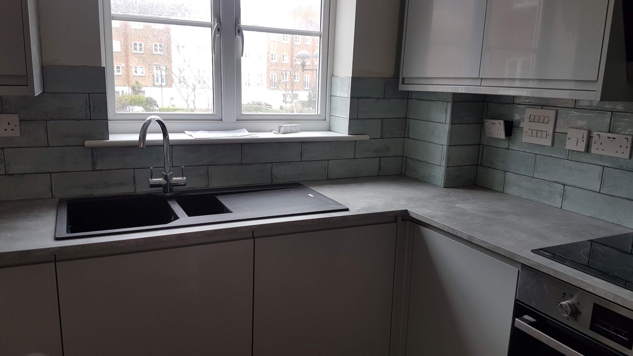 Nowoczesna kuchnia po remoncie: szare szafki, blat imitujący beton, turkusowe płytki na ścianie, okno z widokiem na budynki. Bateria i zlew w minimalistycznym stylu.