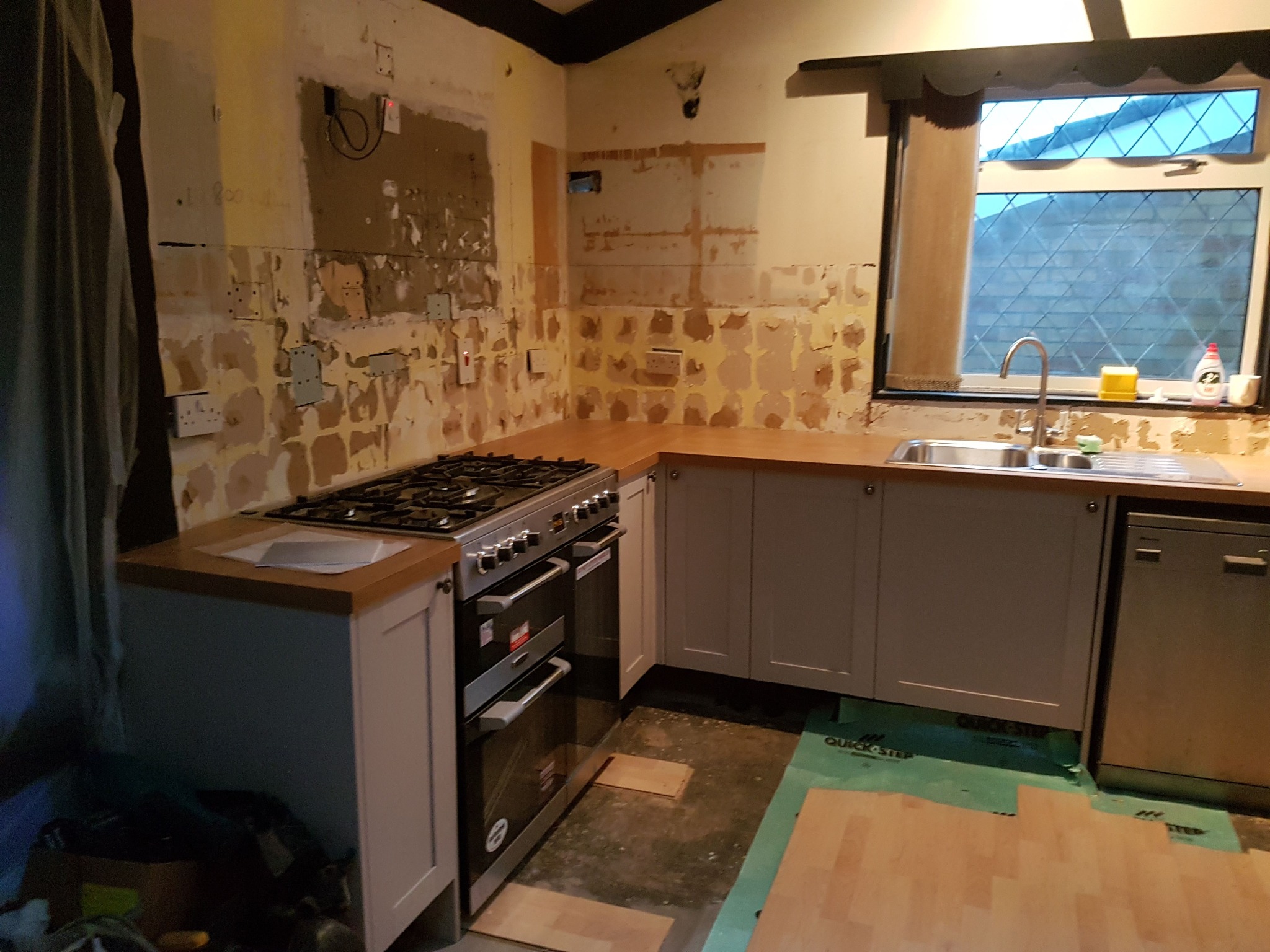 Kuchnia w trakcie remontu: częściowo zamontowane szare szafki z drewnianym blatem, płyta gazowa, fragment ułożonych paneli podłogowych, ściany przygotowane do malowania.