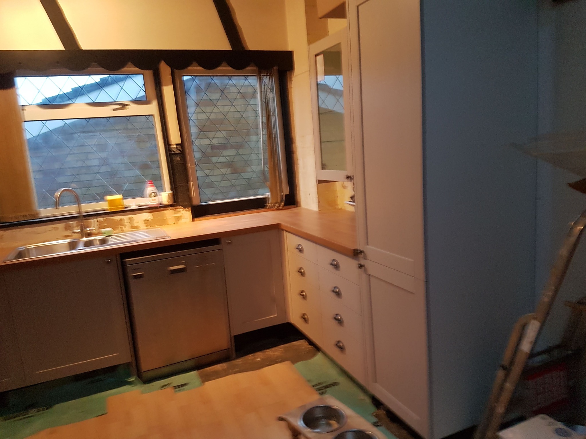 Białe szafki kuchenne z drewnianym blatem w Jazgarzewszczyźnie. Widoczny zlew, okno z szybami w romby i drabina. Częściowo ułożona podłoga.