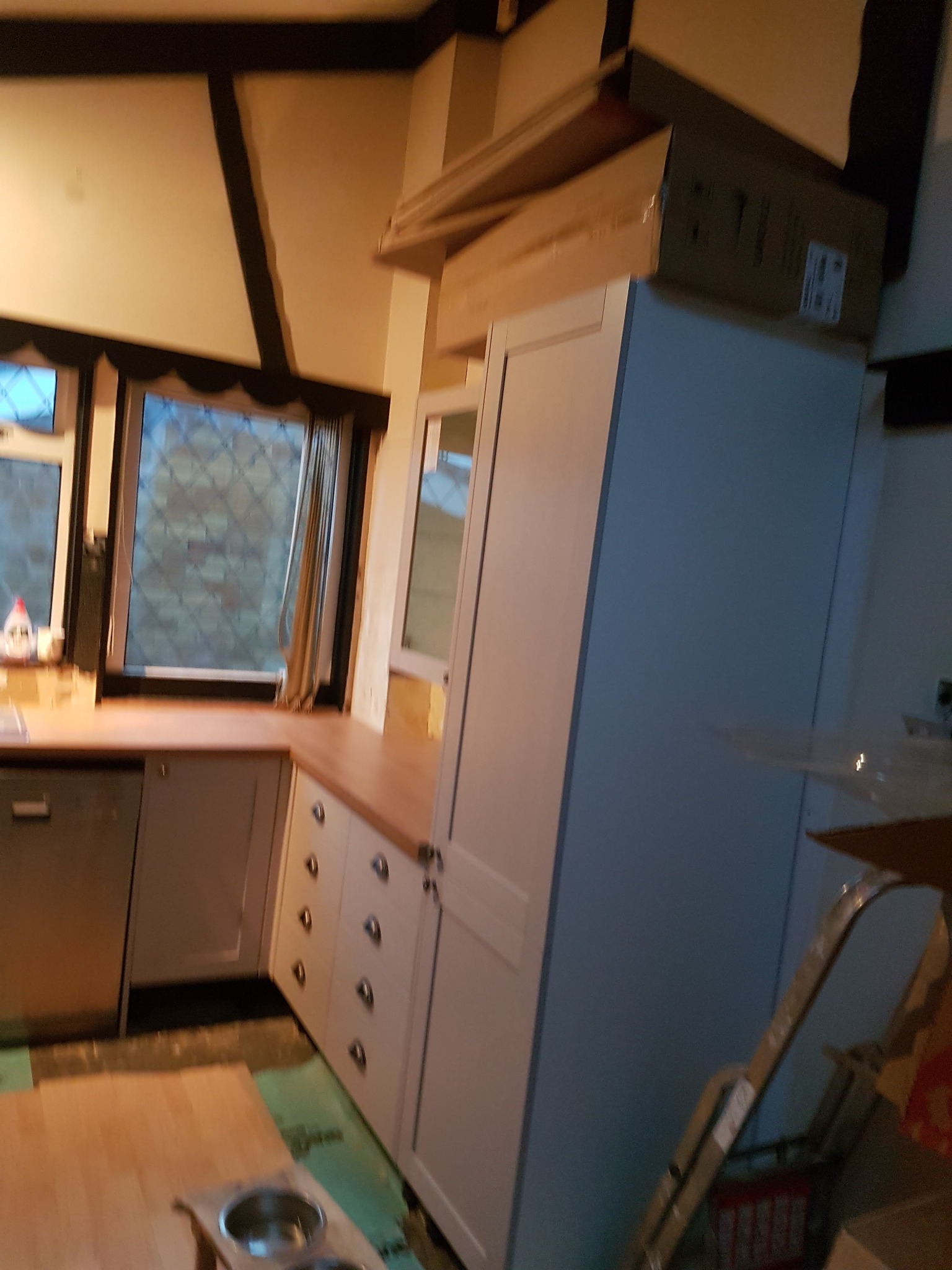 Jasna kuchnia w trakcie montażu: białe szafki z srebrnymi uchwytami, drewniany blat, okno z firanką. Na podłodze widoczne materiały montażowe i drabina.