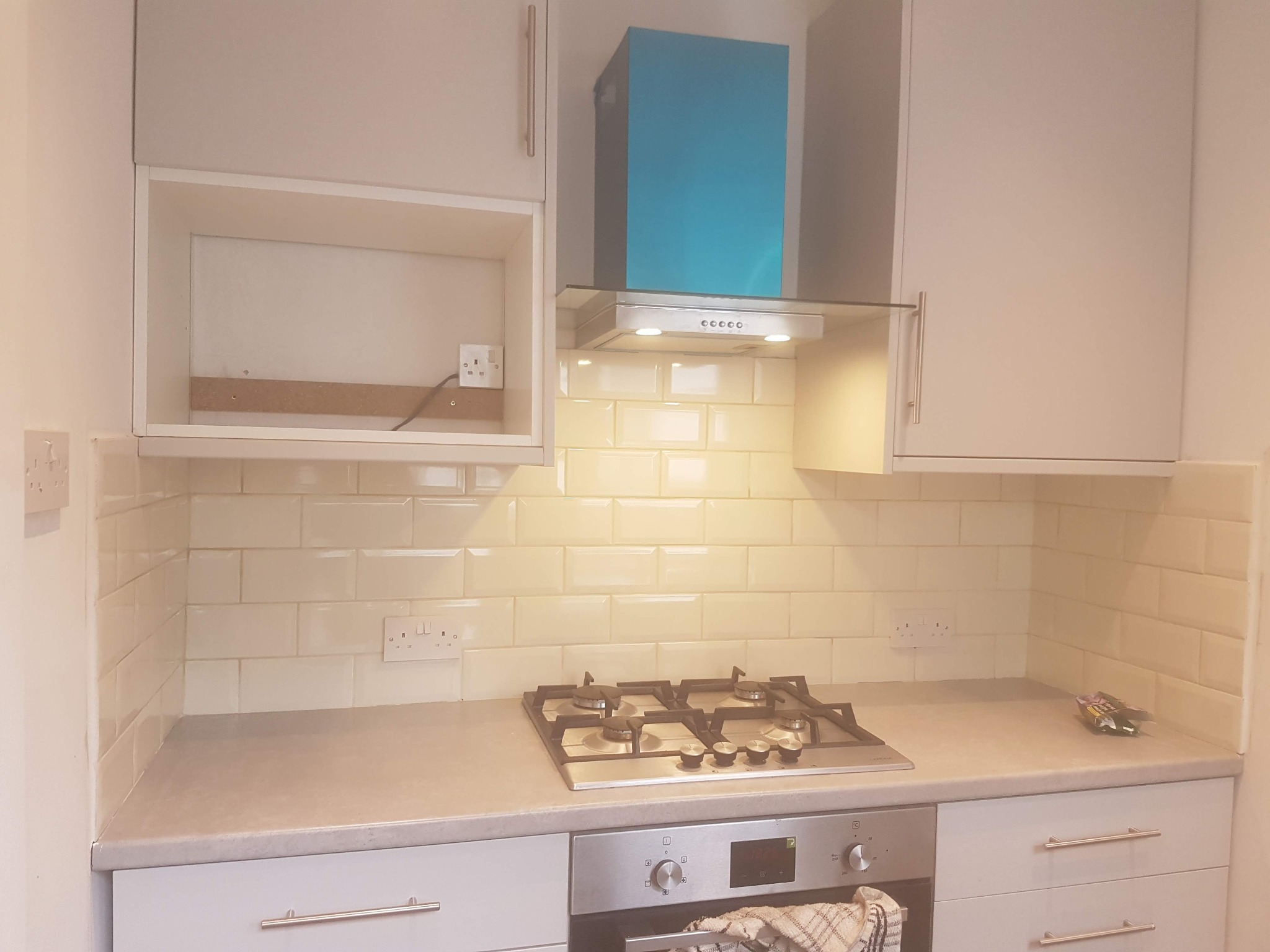 Jasna kuchnia po remoncie: kremowe płytki, białe szafki, srebrny okap i płyta gazowa. Widoczne gniazdka elektryczne i fragment blatu. Ukończony projekt.