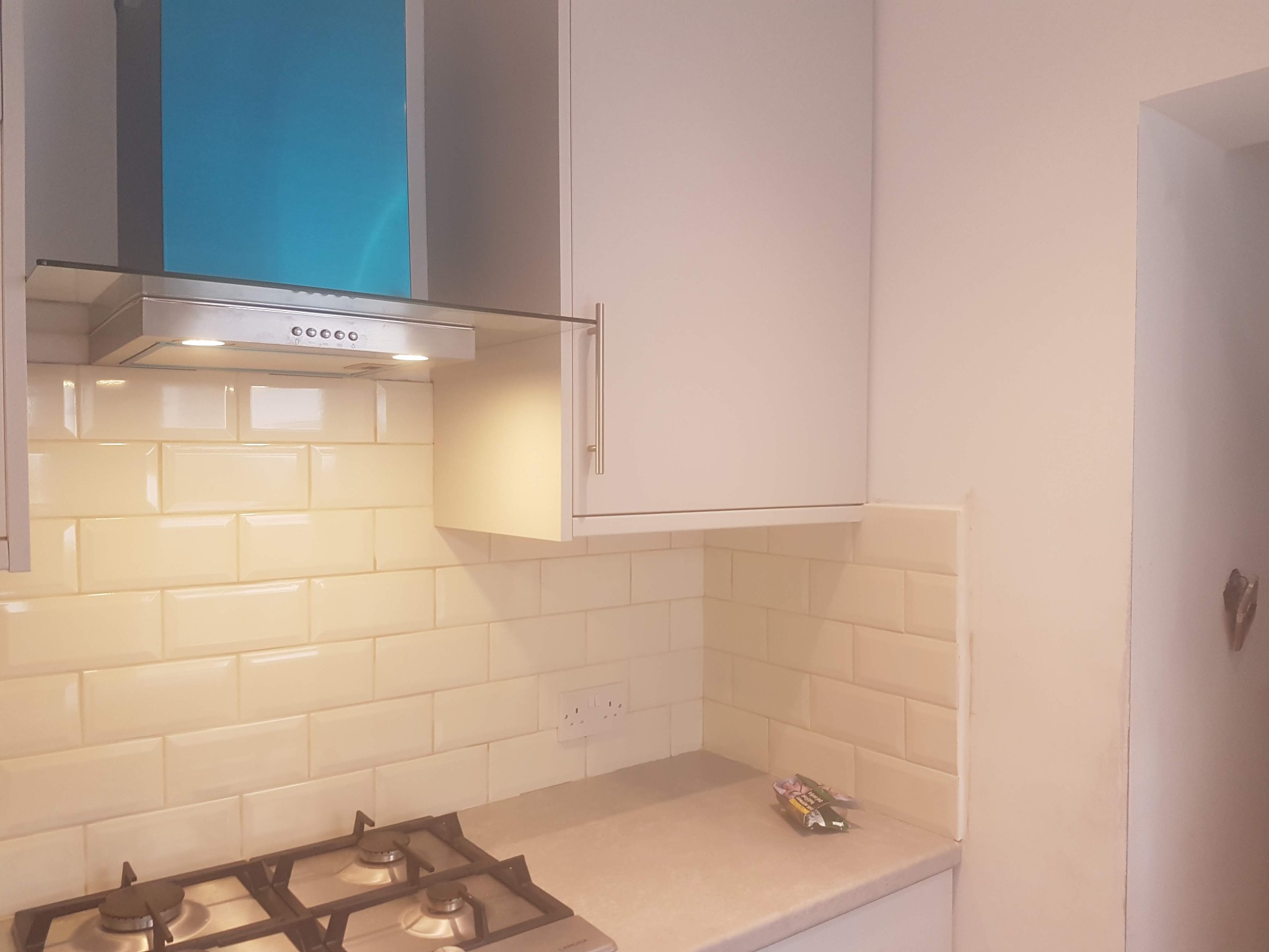 Wykończona kuchnia z beżowymi płytkami na ścianie, srebrnym okapem i jasnymi szafkami. Widoczna płyta gazowa i gniazdko elektryczne. Ujęcie z bliska.