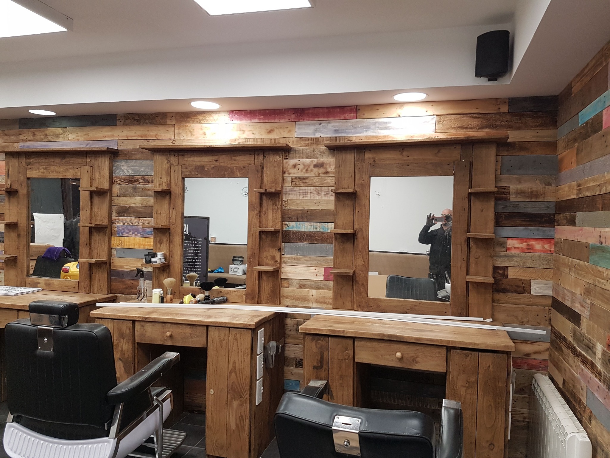 Barbershop z drewnianymi panelami ściennymi i stanowiskami z lustrami. Widoczne fotele fryzjerskie i oświetlenie sufitowe. Unikalny wystrój wnętrza.