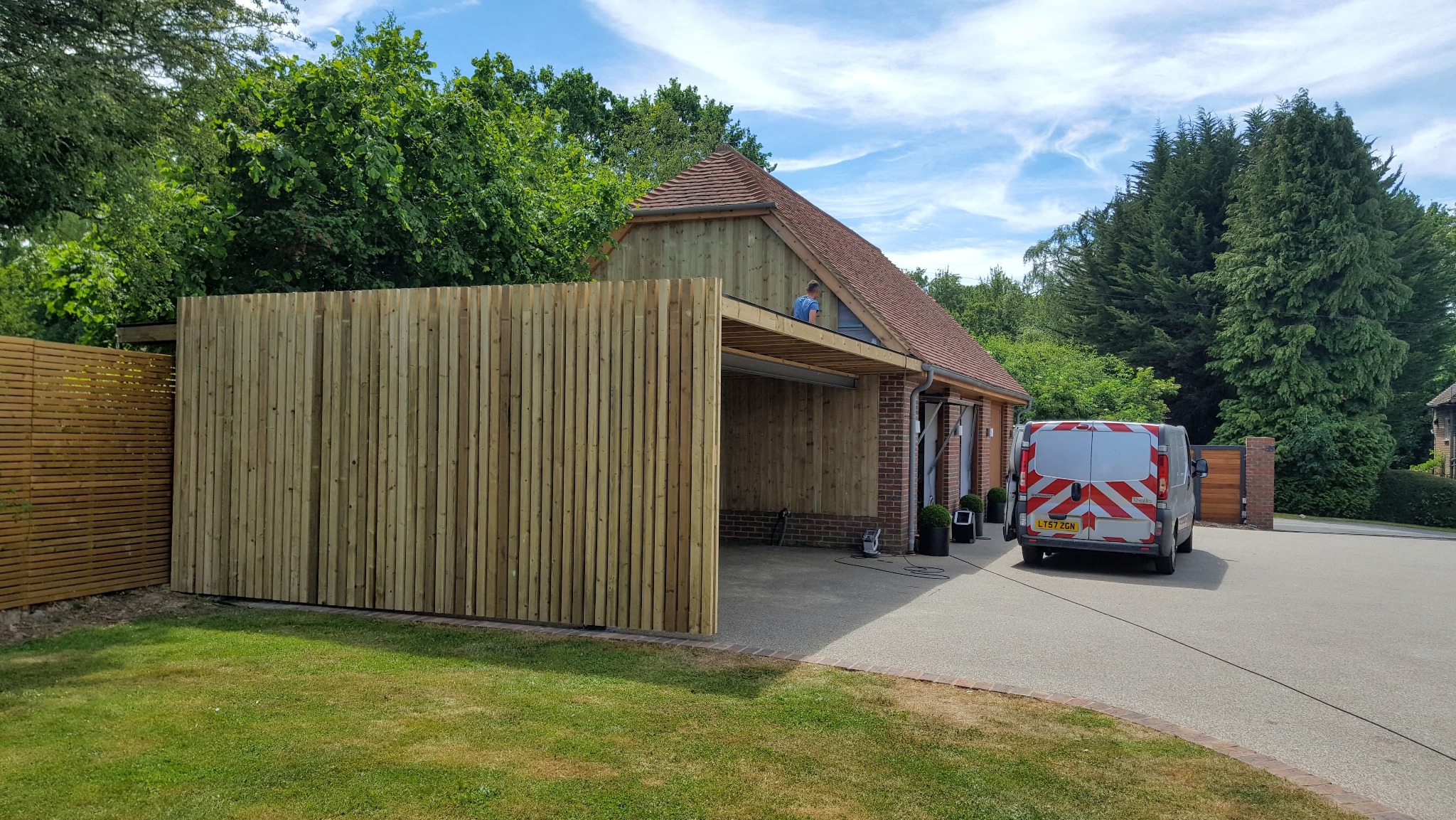 Drewniana konstrukcja w ogrodzie z pionowymi panelami i częściowo zadaszonym miejscem parkingowym, widoczny pracownik na dachu, samochód dostawczy w tle.