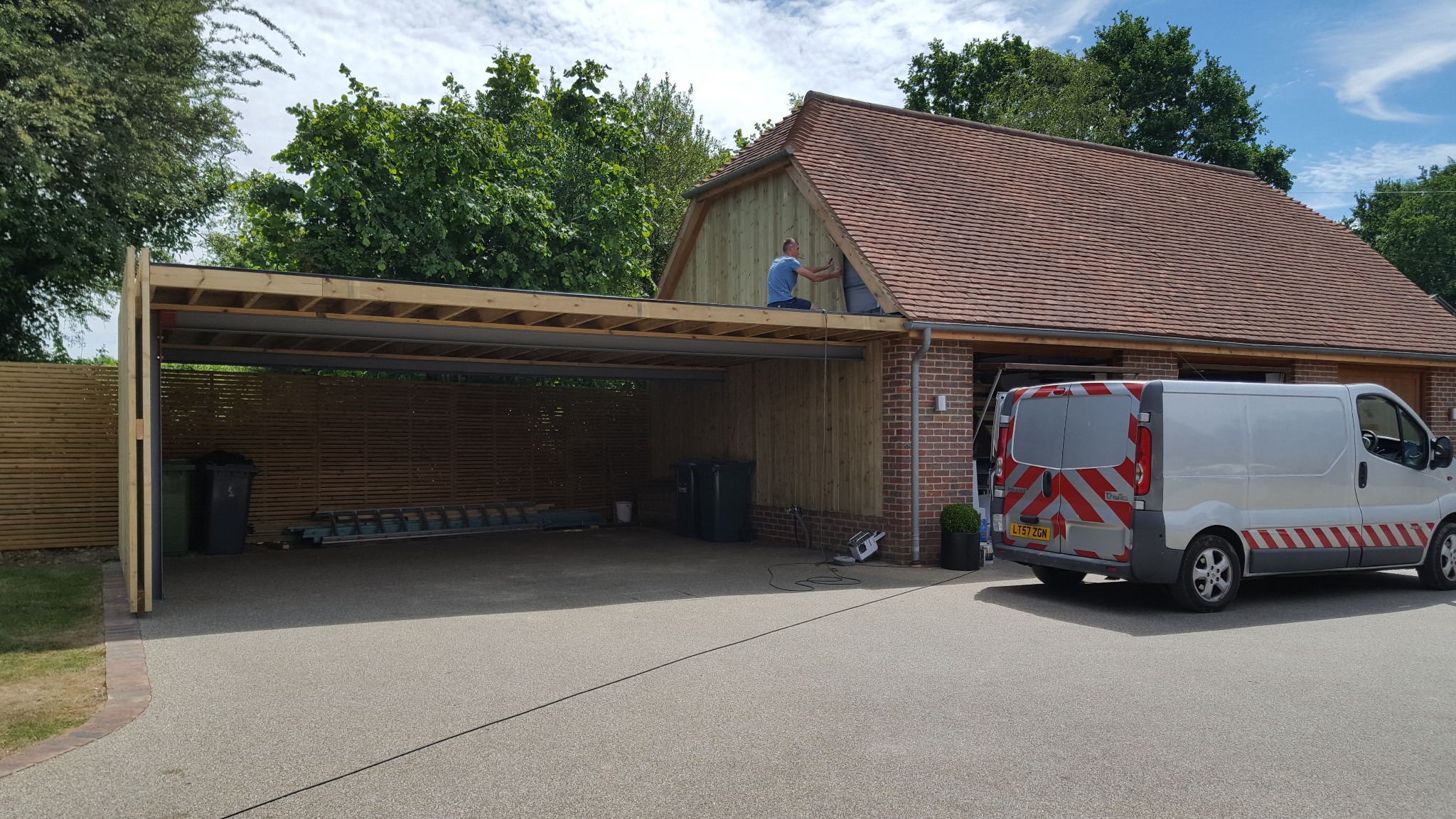 Konstrukcja zadaszenia garażowego w trakcie budowy, widoczny pracownik na dachu, drewniane elementy i ceglana ściana budynku w tle, zaparkowany samochód dostawczy.