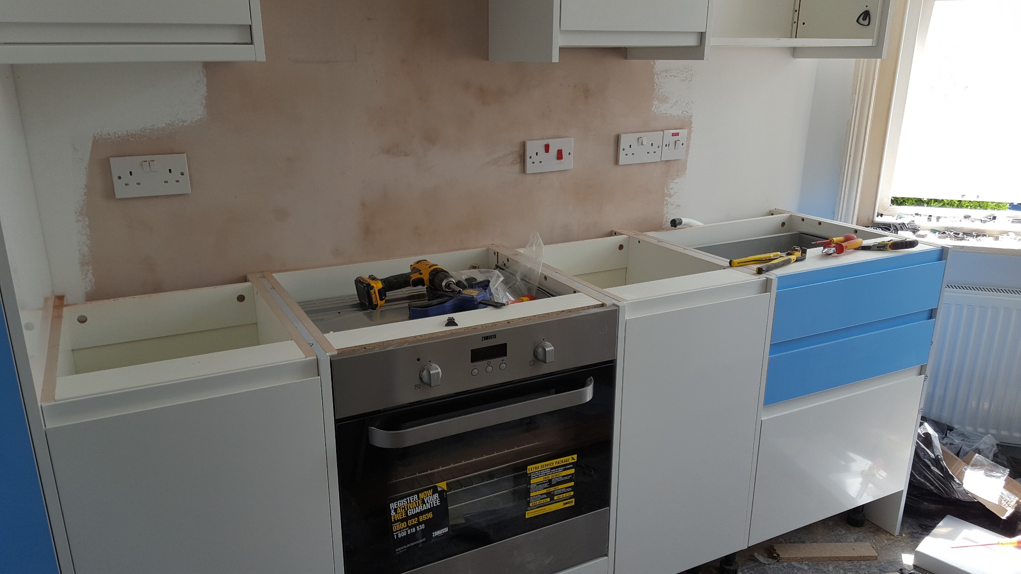 Montaż białych szafek kuchennych z niebieskimi szufladami, widoczne narzędzia i niedokończone ściany. Etap instalacji piekarnika Zanussi.