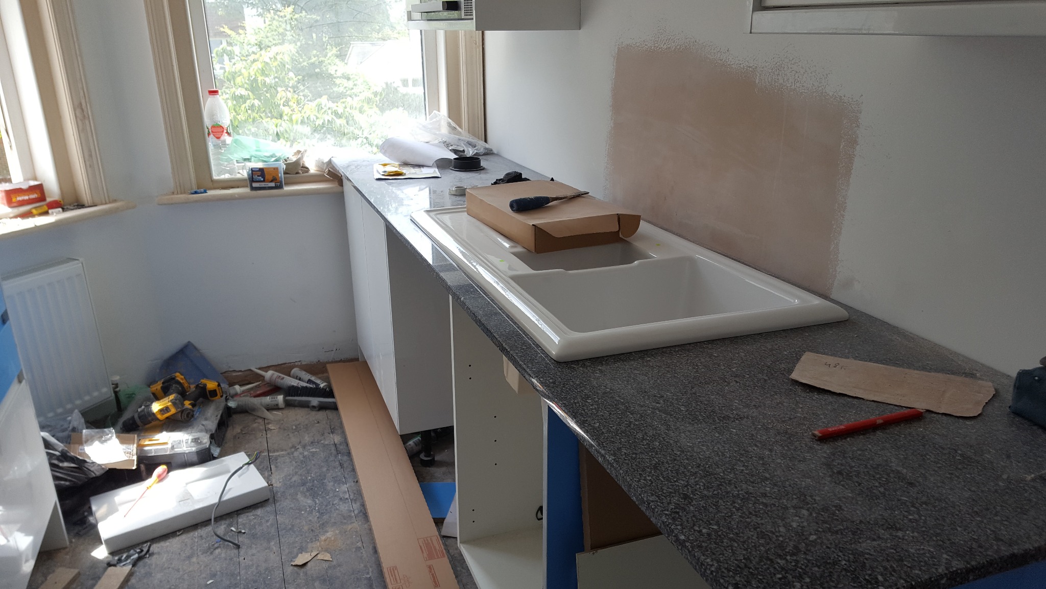 Montaż zlewu w kuchni podczas remontu. Widoczne narzędzia, niedokończone ściany i elementy szafek. Blat z ciemnego kamienia.