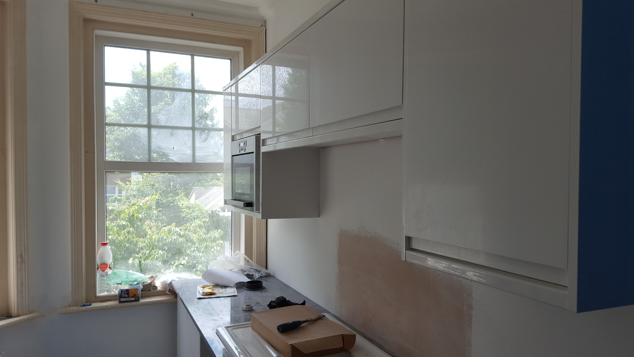 Białe, lakierowane szafki kuchenne z piekarnikiem w zabudowie, widok z boku. Fragment ściany z niedomalowanym fragmentem. Okno z widokiem na zieleń w tle.