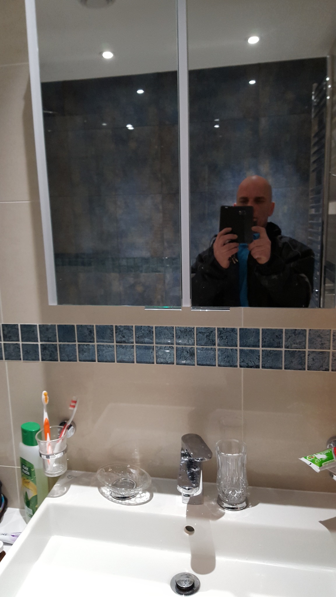 Łazienka z umywalką, baterią i lustrem, w którym odbija się osoba robiąca zdjęcie telefonem. Widoczne płytki dekoracyjne i akcesoria łazienkowe.