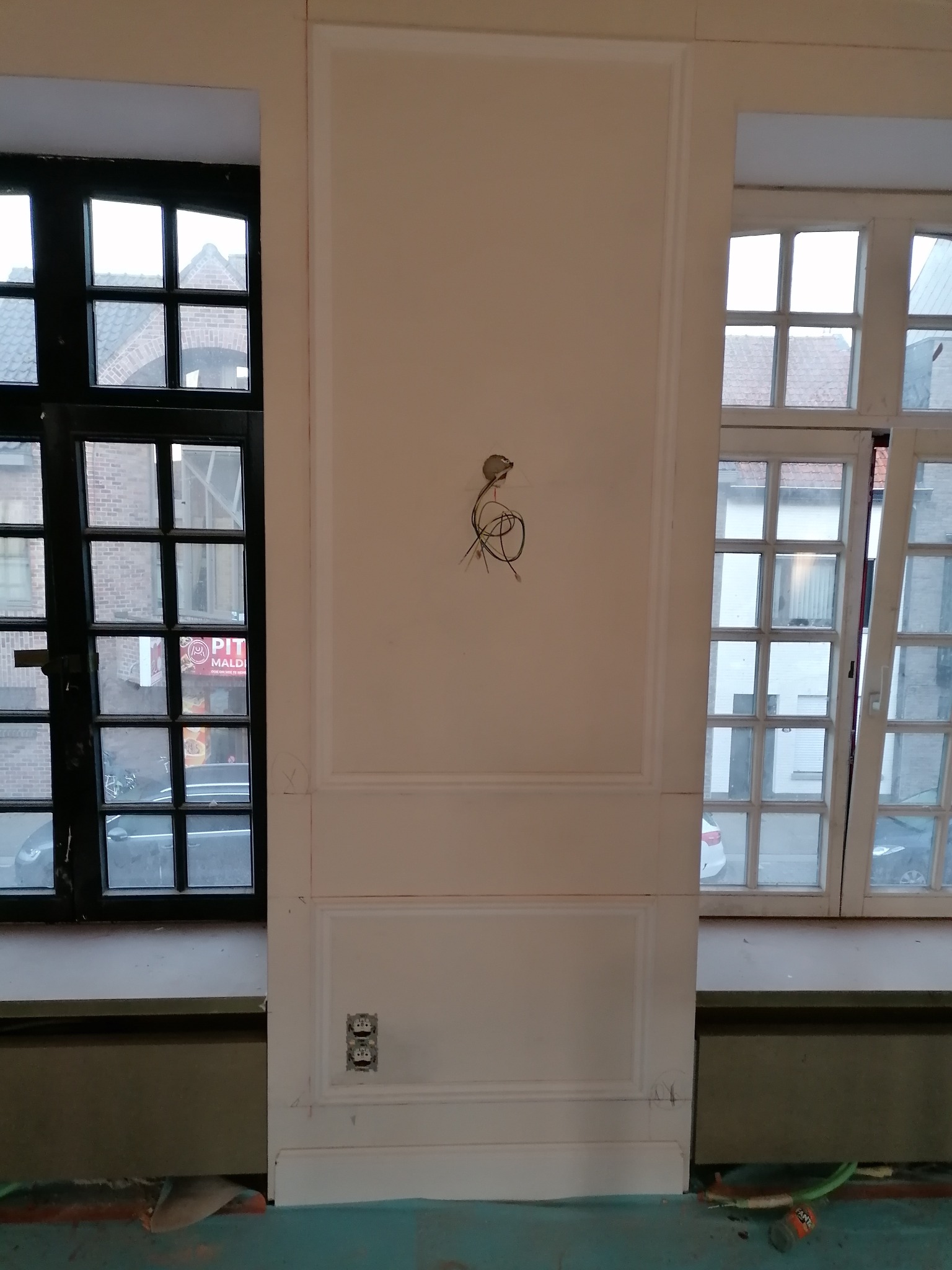 Ściana z dekoracyjnymi panelami, gniazdkami i wystającymi kablami elektrycznymi, pomiędzy dwoma oknami o różnej kolorystyce ram, w trakcie prac wykończeniowych.