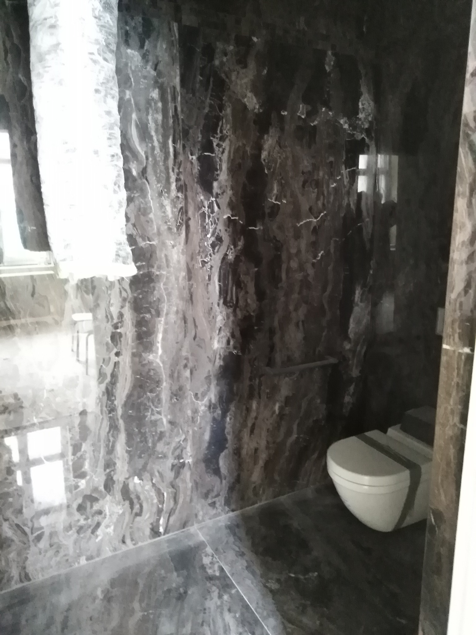 Elegancka łazienka wykończona ciemnym marmurem z wyraźnym użyleniem, widoczna toaleta i fragment okna z zasłoną. Nowoczesny design z akcentem luksusu.