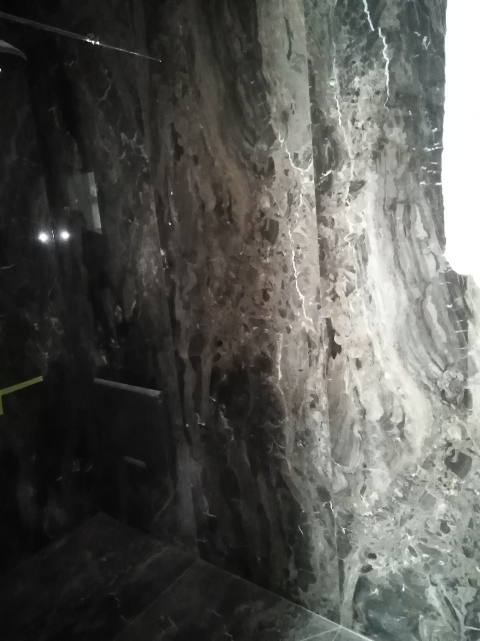 Ciemna, polerowana ściana z efektem marmuru w łazience, z widocznym oświetleniem i odbiciami, ukazująca nowoczesne wykończenie wnętrza.