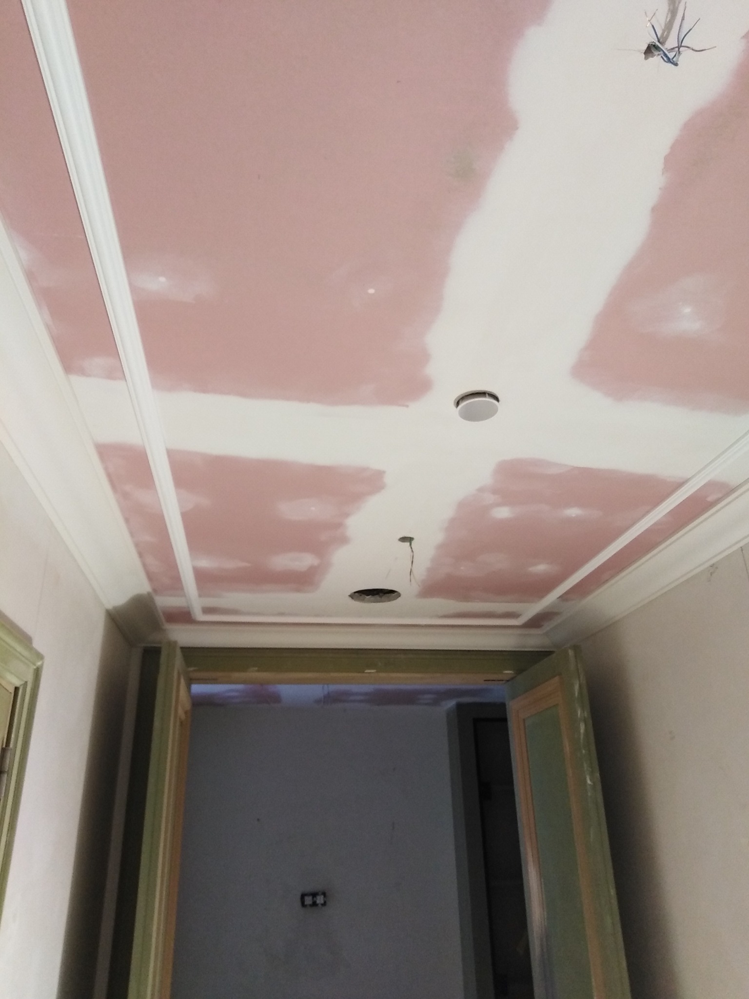 Sufit z płyt gipsowo-kartonowych, częściowo pokryty szpachlą, z dekoracyjnymi listwami i otworami na oświetlenie, widok z dołu na korytarz z drzwiami.