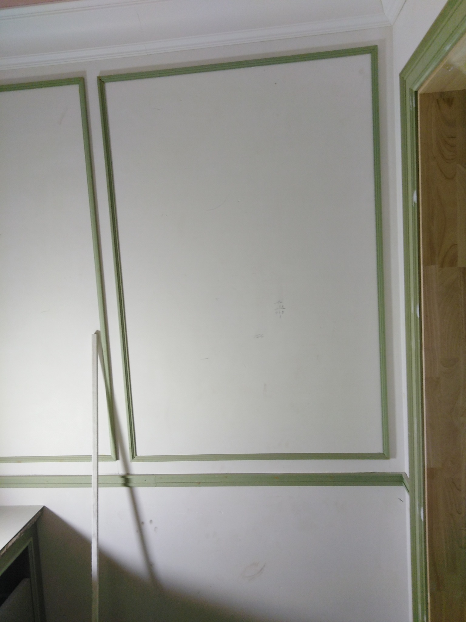 Ściana z białymi panelami i zielonymi listwami dekoracyjnymi. Fragment drabiny oparty o ścianę. Widoczny fragment ościeżnicy drzwi.