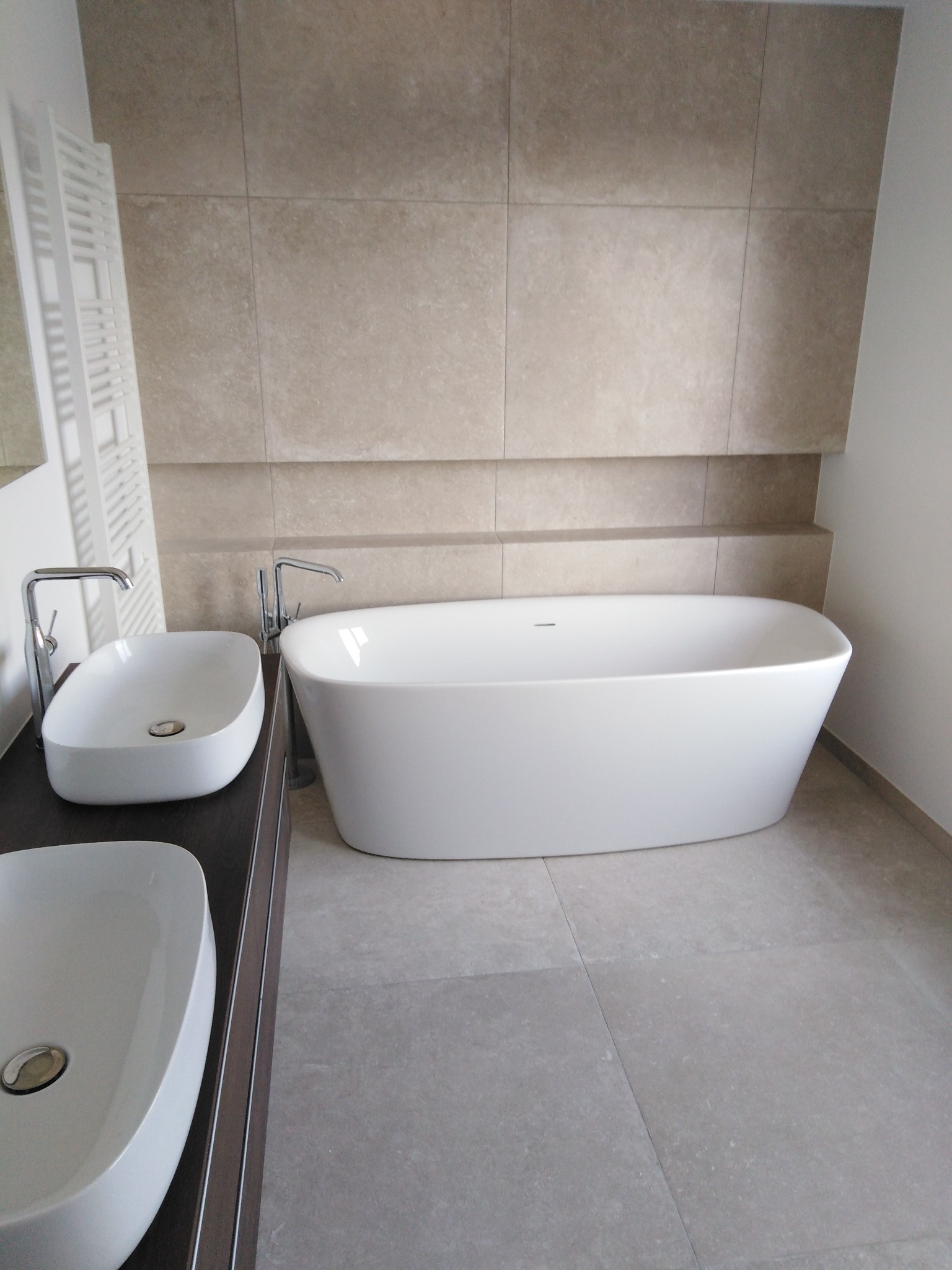 Nowoczesna łazienka z wolnostojącą wanną i dwiema umywalkami nablatowymi. Beżowe płytki na ścianach i podłodze. Minimalistyczny design, chromowana armatura.