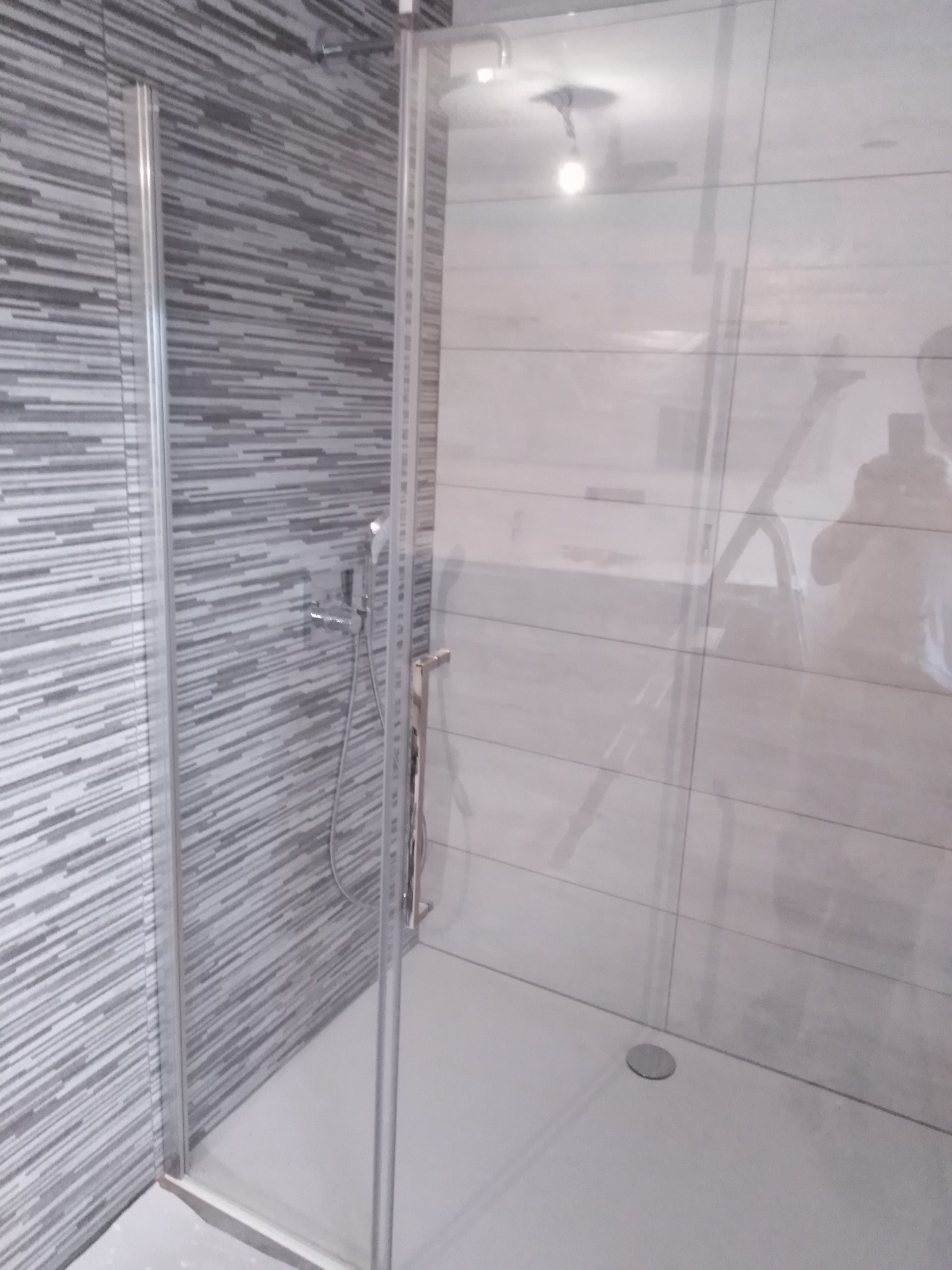 Nowoczesna kabina prysznicowa z szarymi, poziomymi płytkami imitującymi drewno na ścianie. Widoczny chromowany zestaw prysznicowy i szklane drzwi.