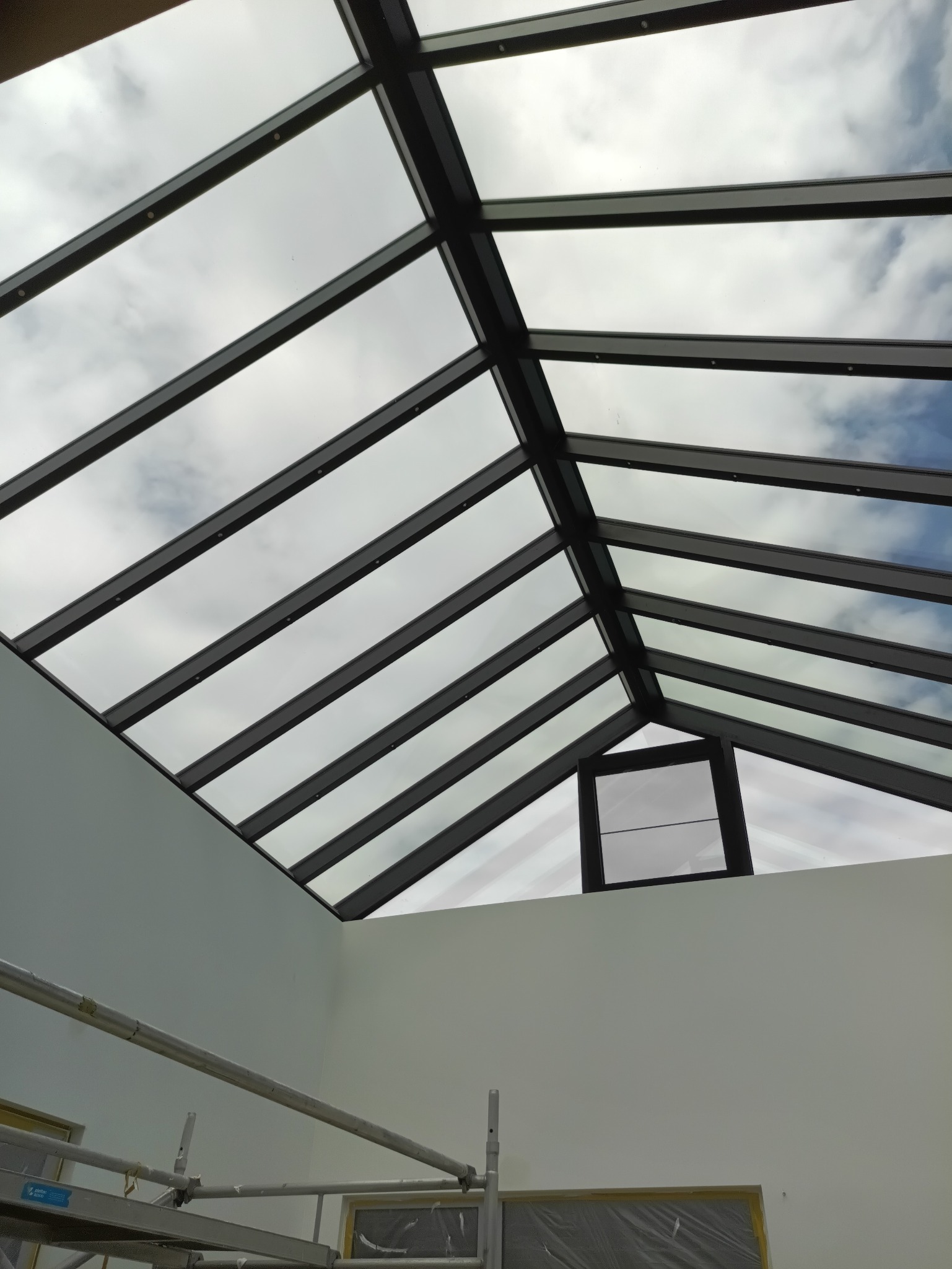 Szklany dach z czarną konstrukcją w jasnym pomieszczeniu, widok z dołu. Fragment rusztowania w lewym dolnym rogu. Widoczne niebo przez szklane panele.