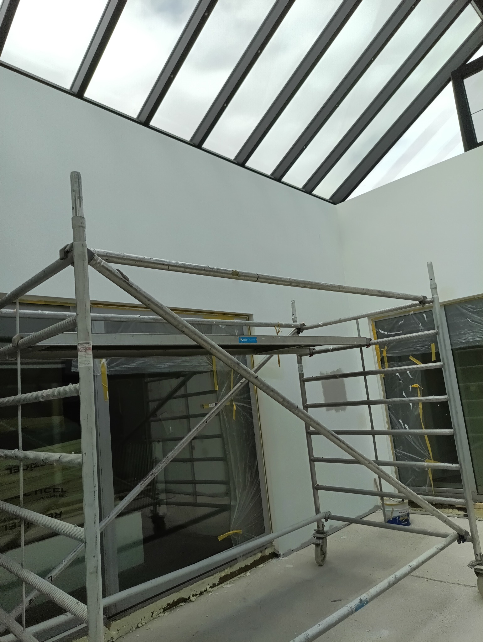 Białe ściany w jasnym wnętrzu z przeszklonym dachem, widoczne rusztowanie malarskie i zabezpieczone okna folią, sugerujące prace wykończeniowe.