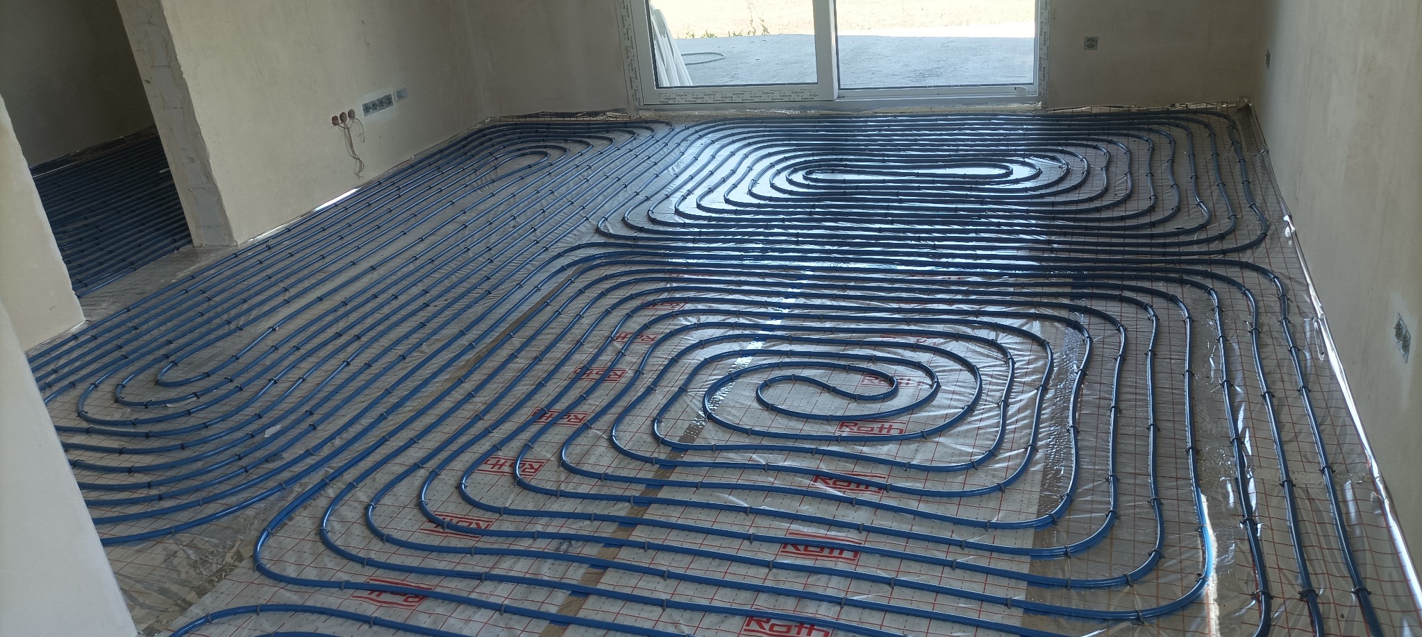 Niebieskie rury ogrzewania podłogowego ułożone spiralnie na folii z siatką w surowym wnętrzu, widok z góry na instalację przed wylaniem posadzki.