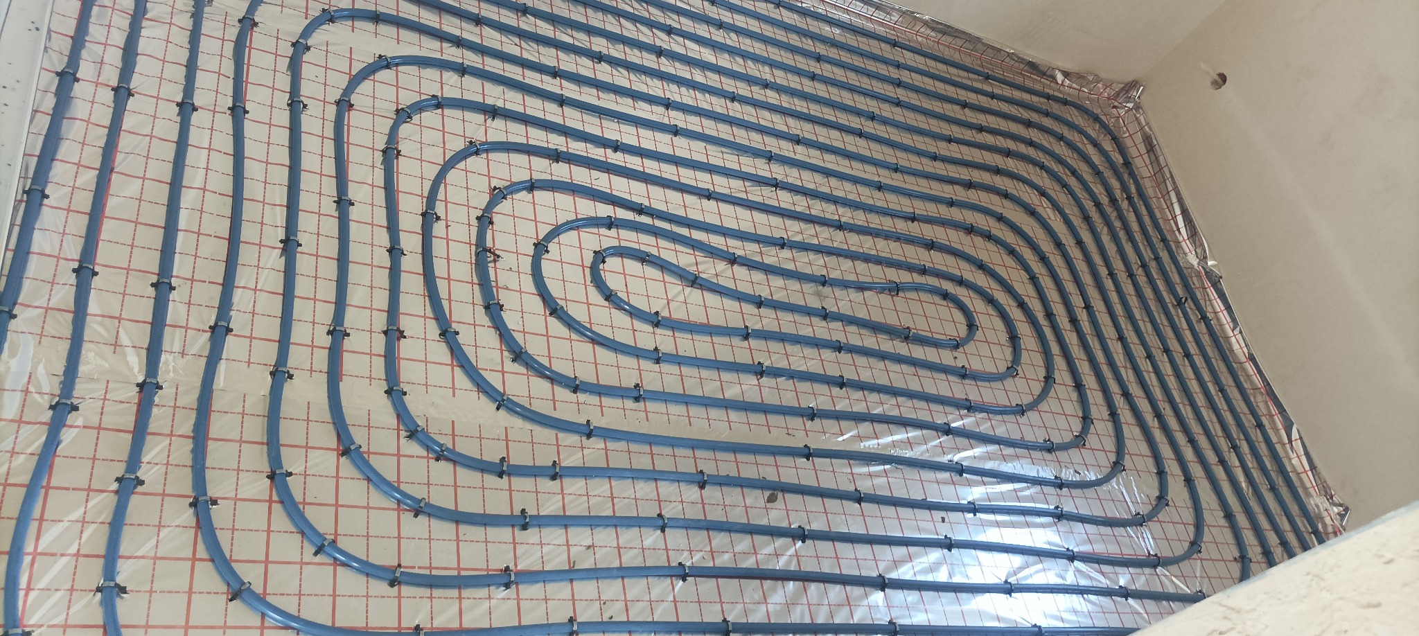 Niebieskie rury ogrzewania podłogowego ułożone spiralnie na folii z czerwoną siatką, przymocowane czarnymi uchwytami. Widok z góry, ukośny kadr.