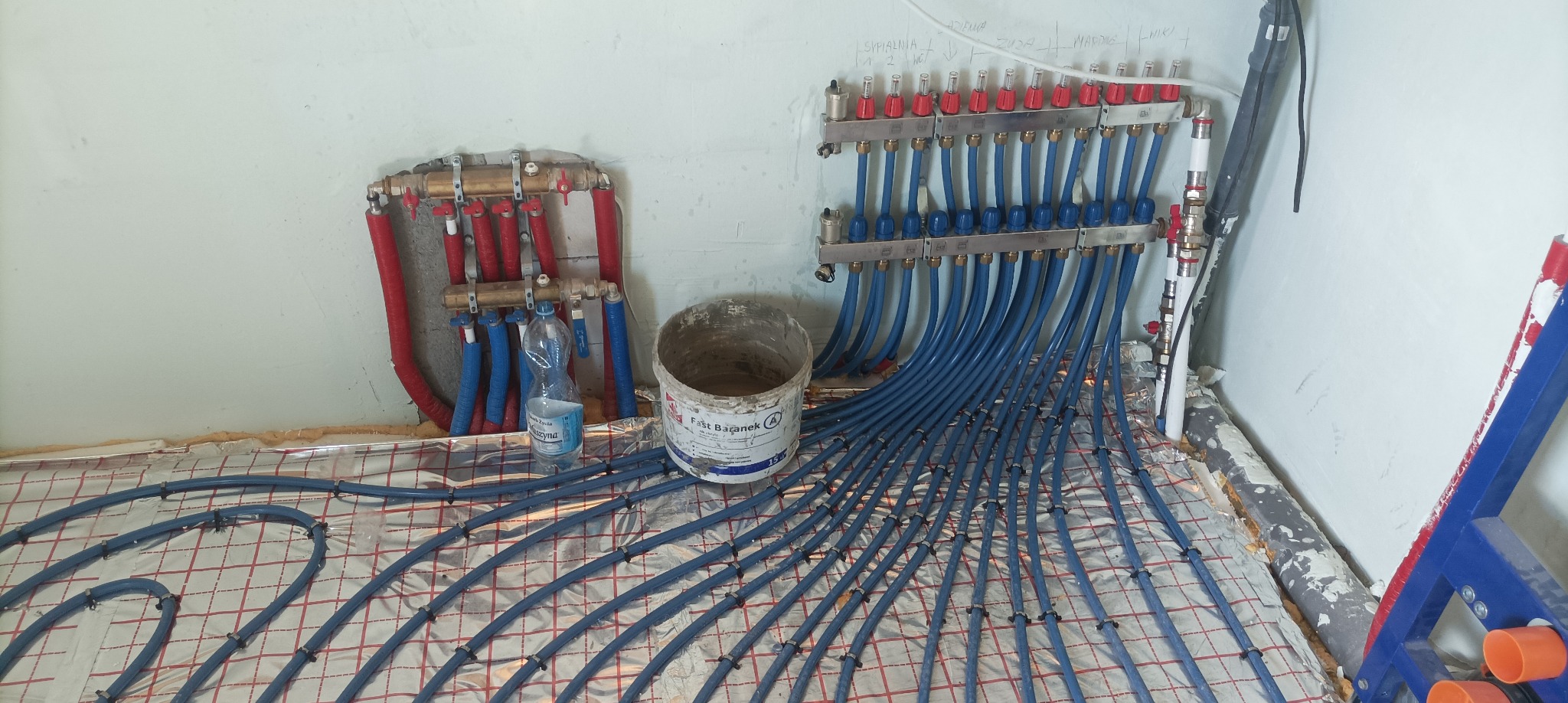 Rozdzielacz i niebieskie rury ogrzewania podłogowego ułożone na folii izolacyjnej z siatką. Widoczny fragment ściany i drabina malarska.