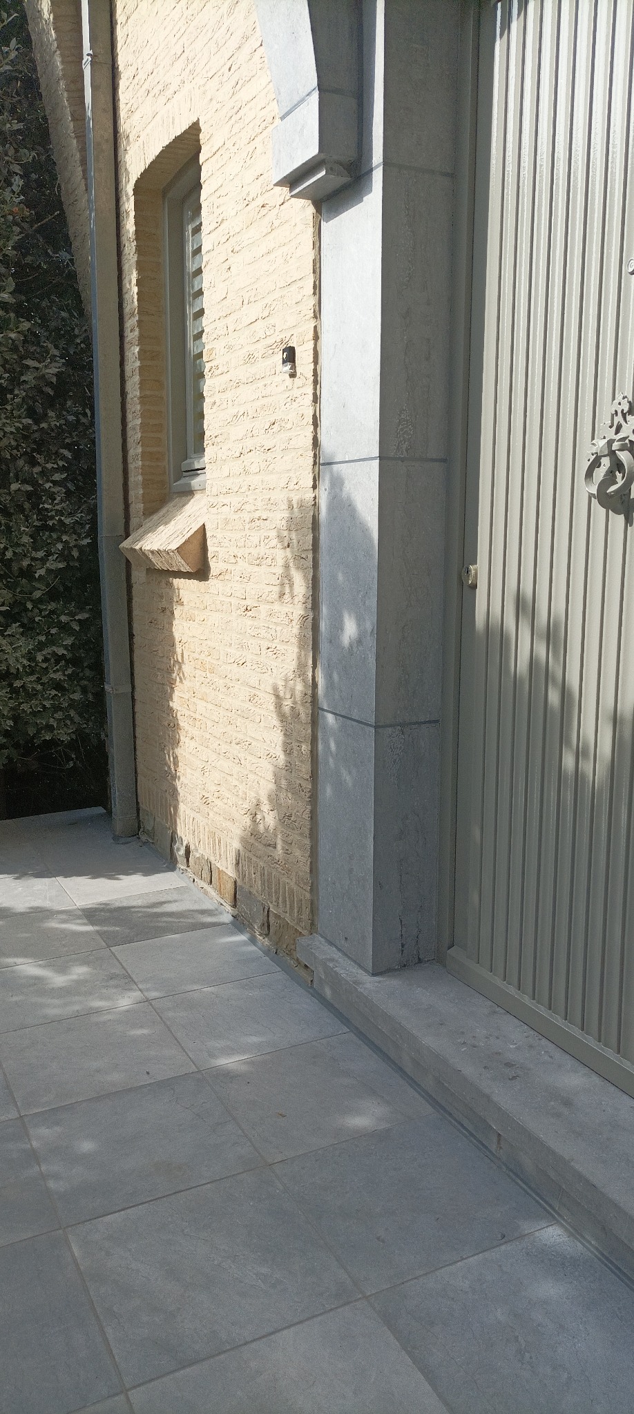 Elewacja budynku z beżowej cegły z szarą, metalową bramą i kamienną kolumną. Widoczne szare płytki chodnikowe i cień drzewa.