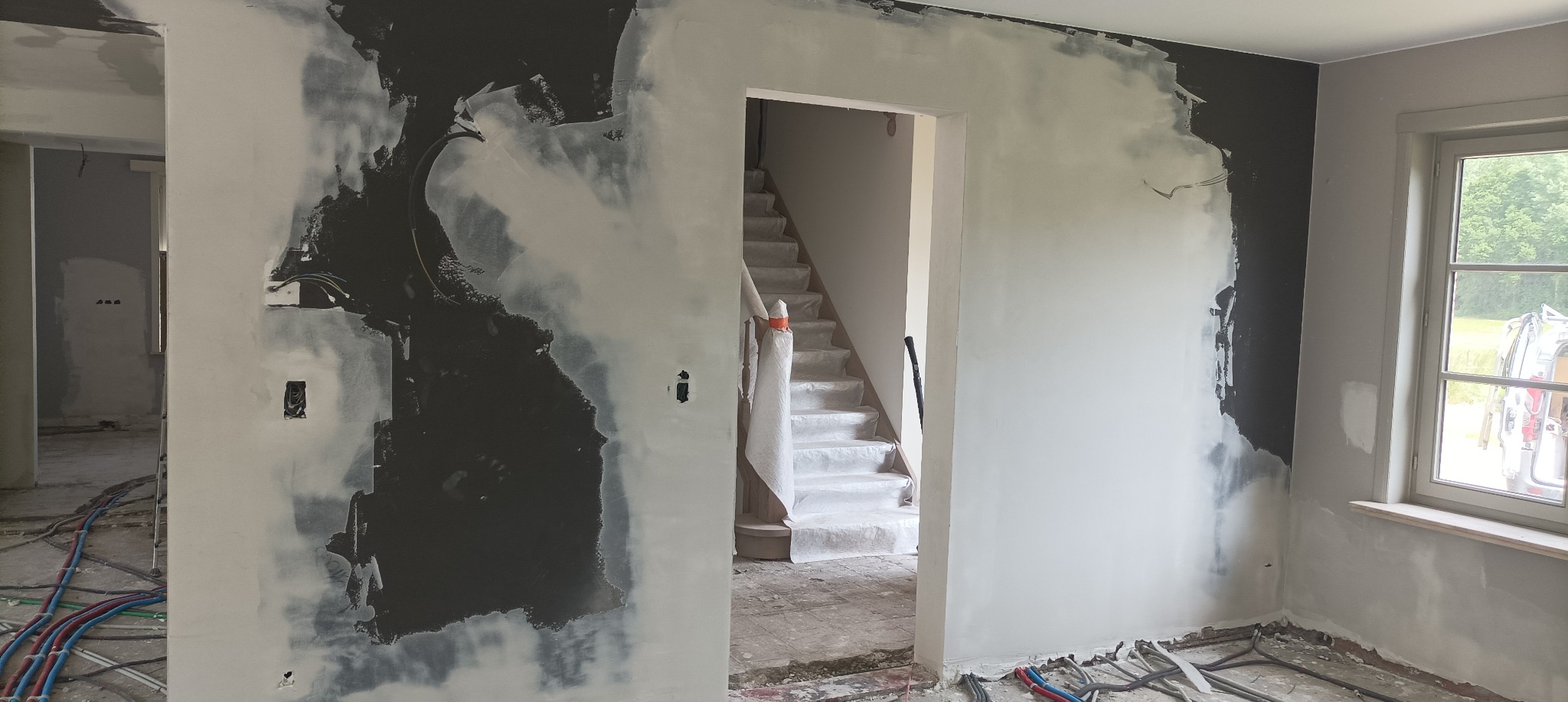 Wnętrze w trakcie prac wykończeniowych: ściany częściowo pokryte szpachlą, widoczne instalacje na podłodze, schody zabezpieczone folią. Okno z widokiem na zieleń.