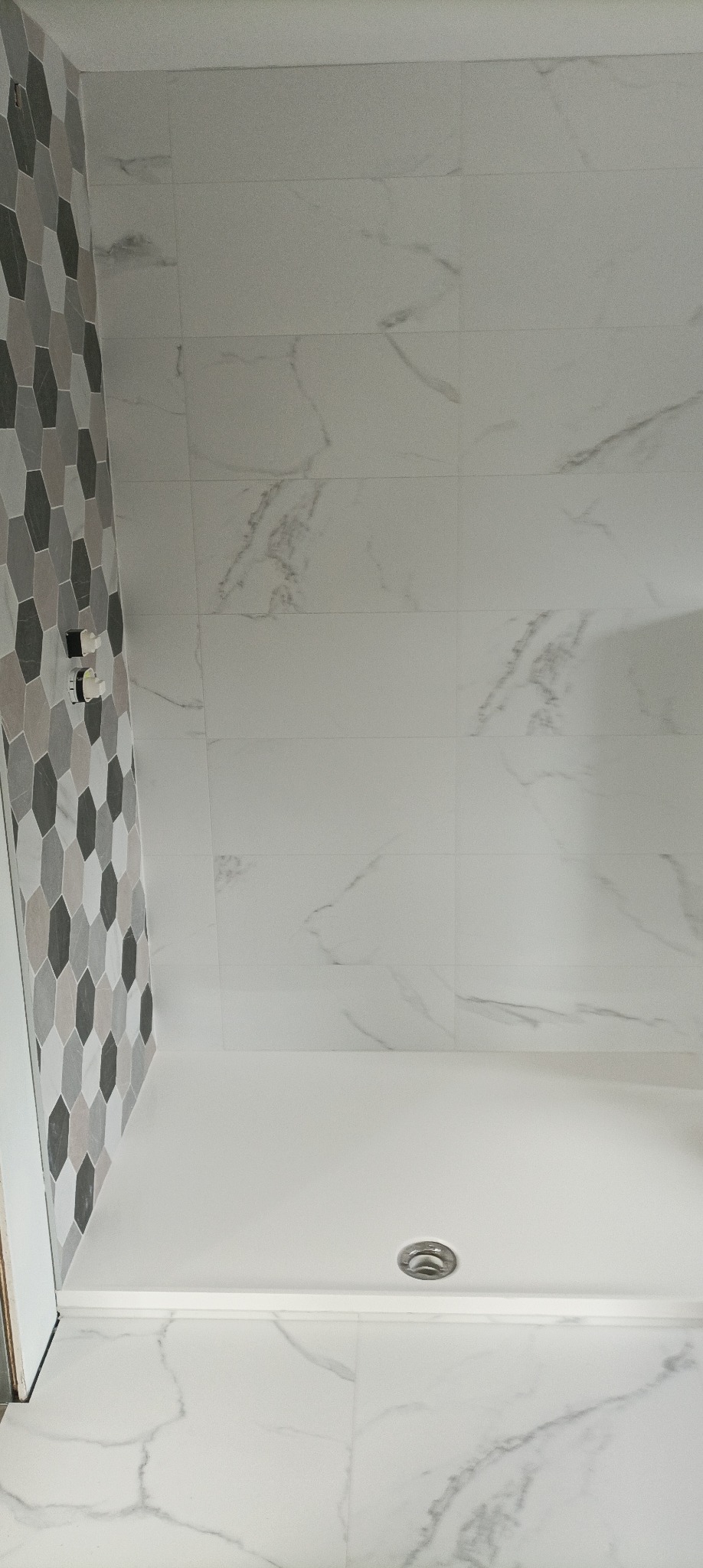 Nowoczesna kabina prysznicowa z białą armaturą i heksagonalną mozaiką w odcieniach szarości. Ściany wyłożone płytkami imitującymi marmur. Minimalistyczny design.
