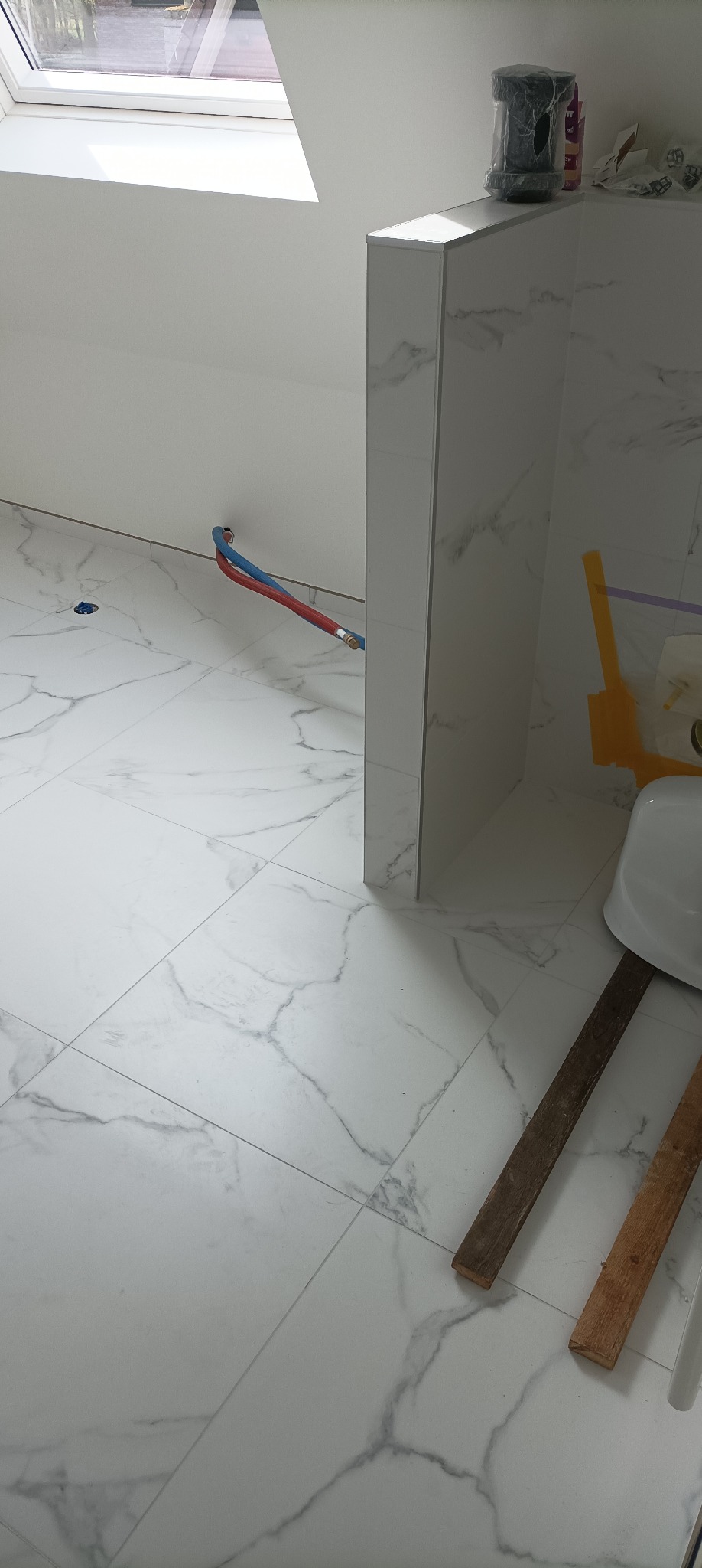 Biała łazienka na poddaszu z marmurową glazurą, widoczne rury instalacji wodnej i fragment umywalki. Ukończone wykończenie podłogi i ścian.