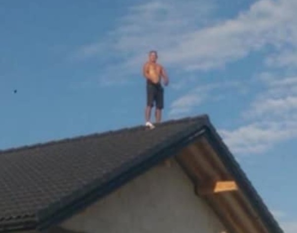 Mężczyzna bez koszulki stoi na ciemnym dachu domu jednorodzinnego na tle błękitnego nieba z chmurami. Ujęcie z dołu.