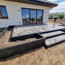 MB-BRUK SAMANTA BORUCZKOWSKA - Nowoczesny taras z kostki brukowej w odcieniach szarości, z niskimi schodami i obrzeżami, na tle białego domu z dużymi oknami. Widoczna osoba w tle i częściowo wypełniona taczka.