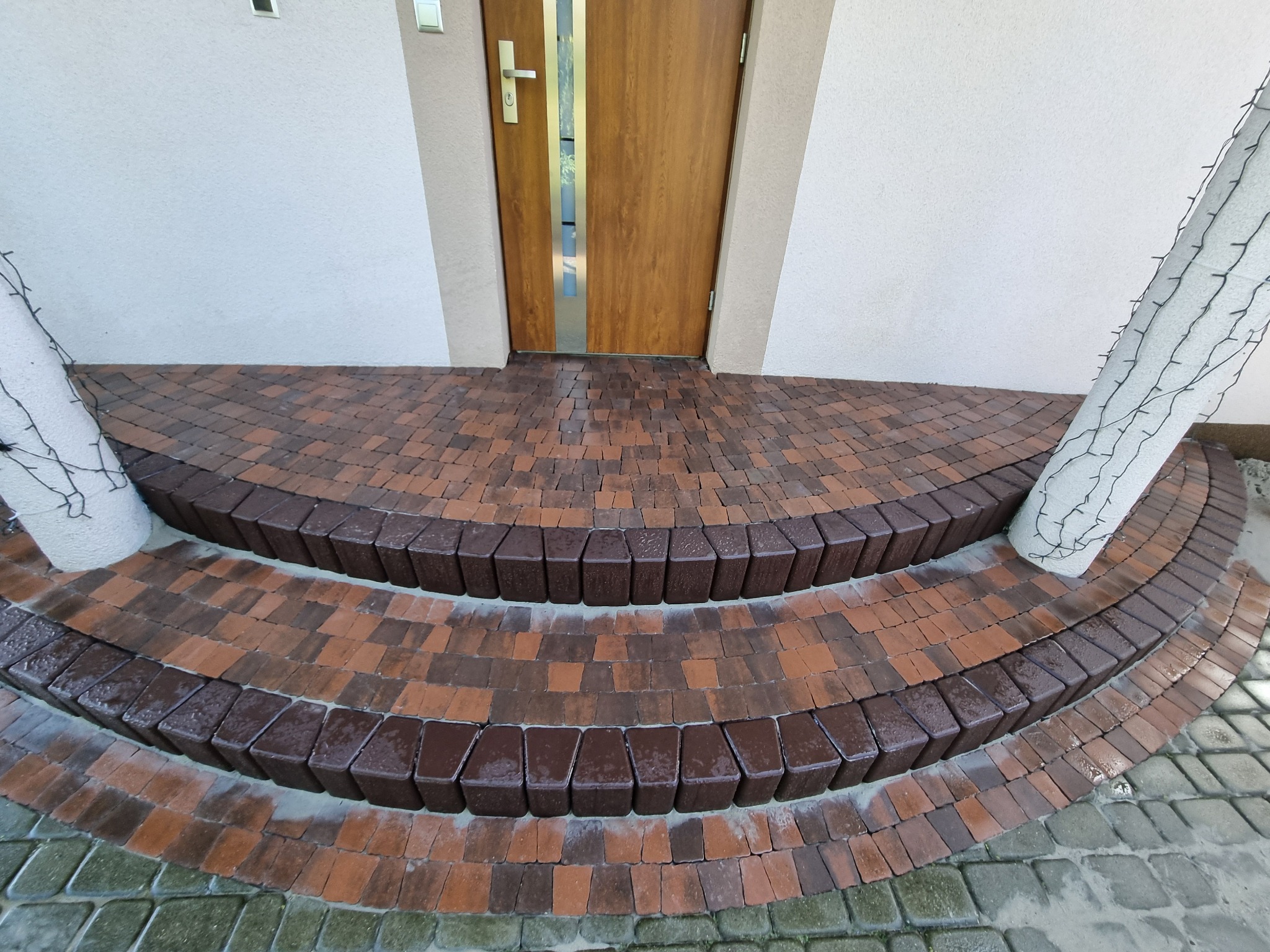 Schody wejściowe z kostki brukowej w odcieniach brązu i beżu, prowadzące do drewnianych drzwi wejściowych. Dekoracyjne oświetlenie na filarach.