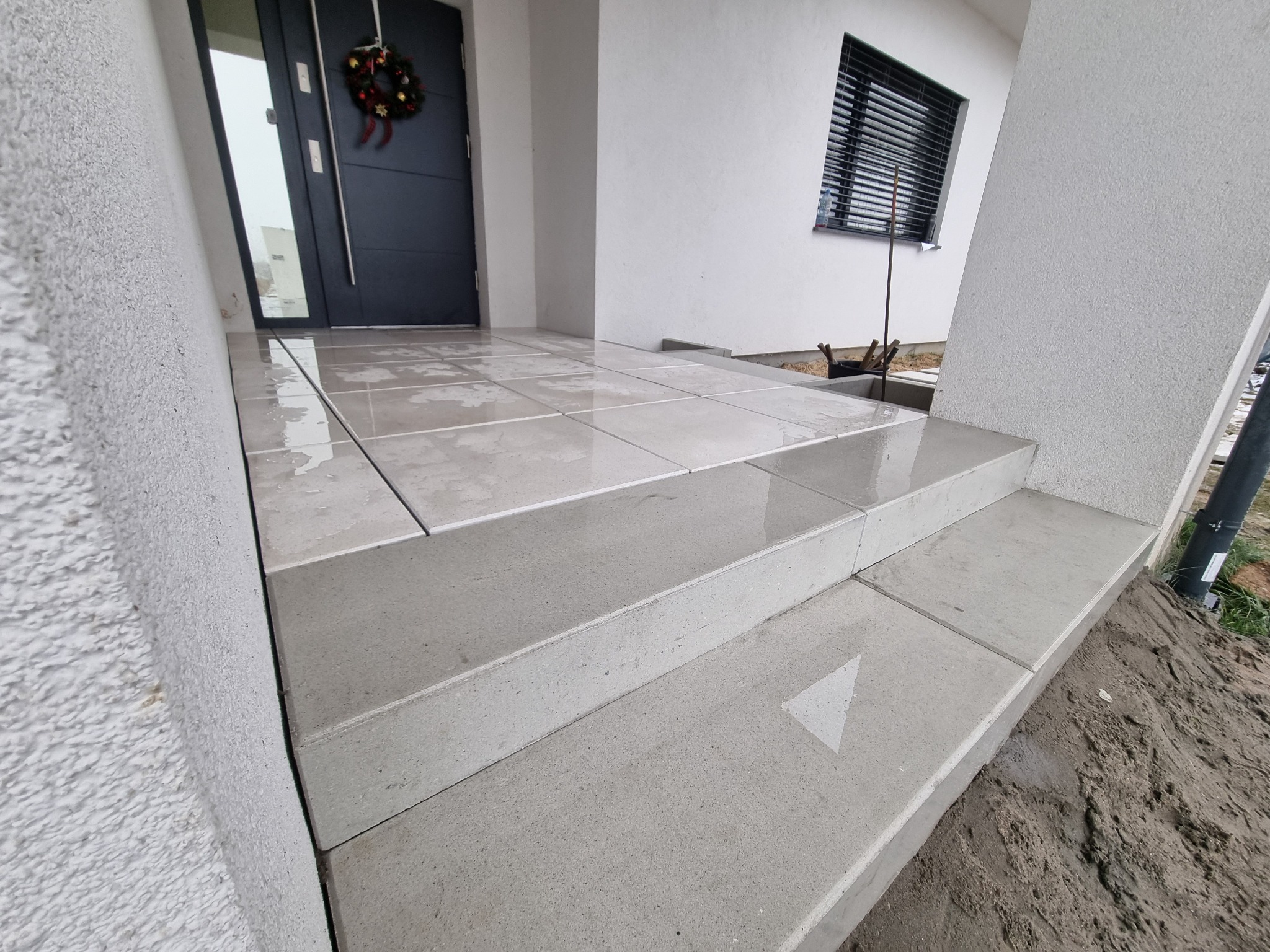 Wykończone schody wejściowe z jasnoszarej kostki brukowej, prowadzące do nowoczesnego domu z ciemnymi drzwiami i świątecznym wieńcem. Widoczne narzędzia brukarskie.