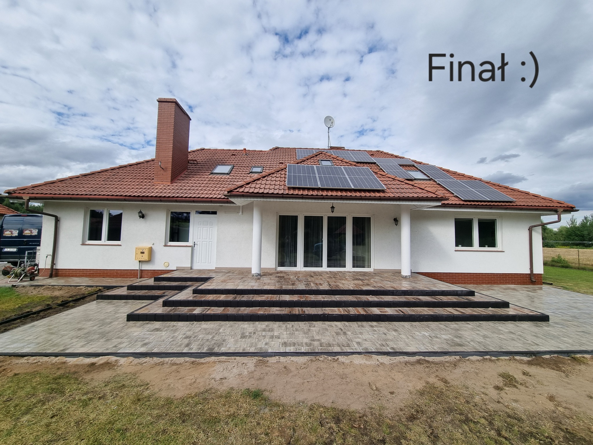 Nowoczesny dom jednorodzinny z tarasem wyłożonym kostką brukową w odcieniach szarości i czerni. Na dachu panele słoneczne. Widoczny komin i fragment ogrodu.