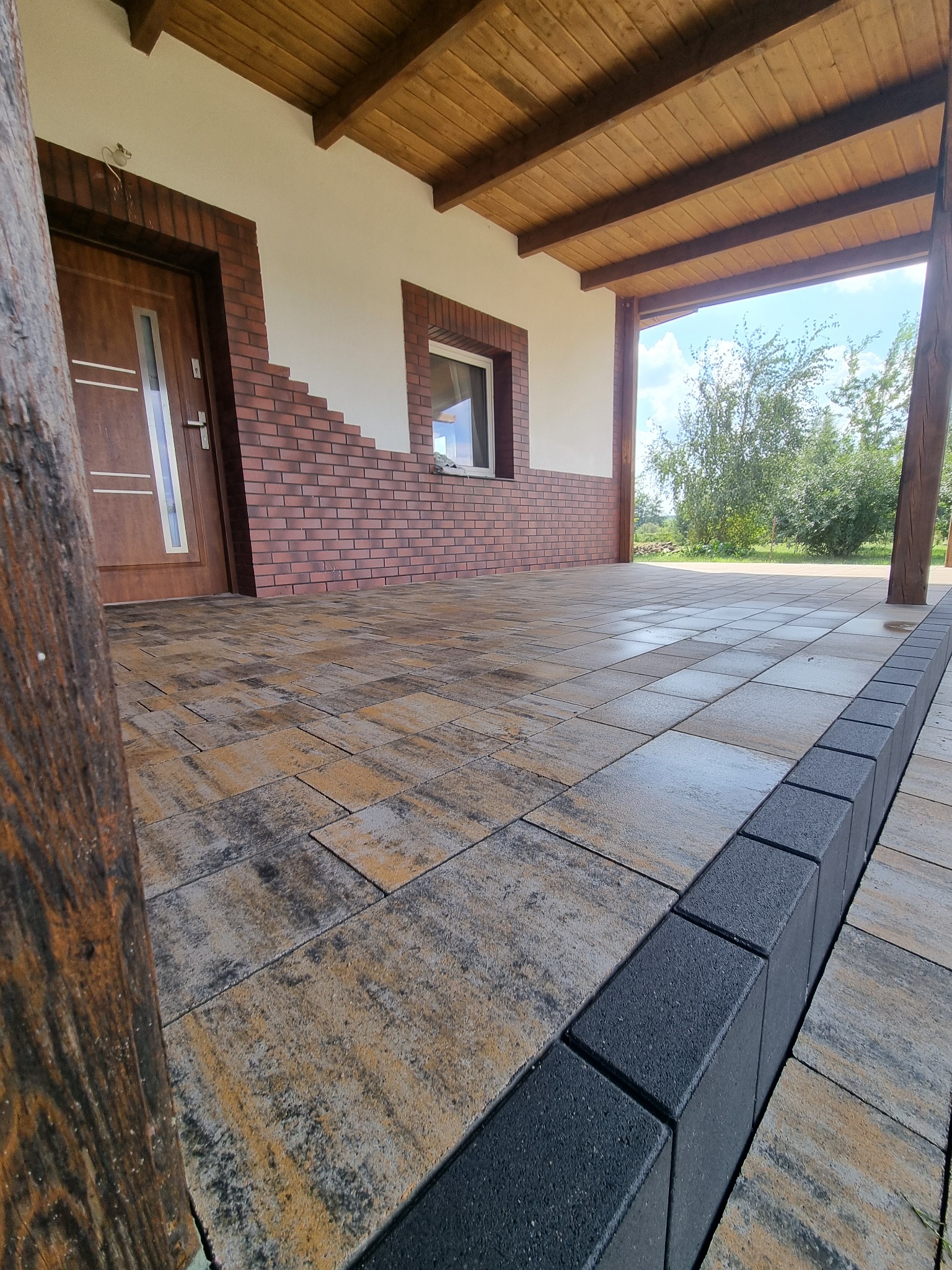 Ułożona kostka brukowa w odcieniach szarości i beżu na zewnątrz budynku z drewnianą kolumną i obrzeżami z ciemnej kostki. Widok z niskiej perspektywy.