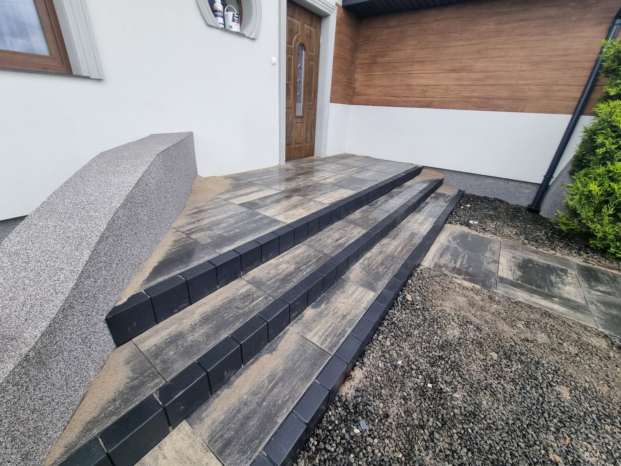 Schody wejściowe z kostki brukowej w odcieniach szarości, z czarnymi obrzeżami, prowadzące do drewnianych drzwi wejściowych. Obok widoczny fragment elewacji budynku.