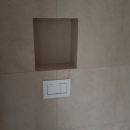 Dariusz Leśniewski L-DAR-BUD - Beżowa, wykafelkowana ściana w łazience z wnęką i przyciskiem spłukującym nad toaletą. Minimalistyczny design, neutralna kolorystyka, nowoczesne wykończenie.