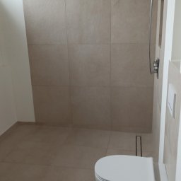 Dariusz Leśniewski L-DAR-BUD - Beżowa łazienka z kabiną typu walk-in i podwieszaną toaletą. Minimalistyczny design, płytki w odcieniach beżu, nowoczesna armatura. Widoczny odpływ liniowy.